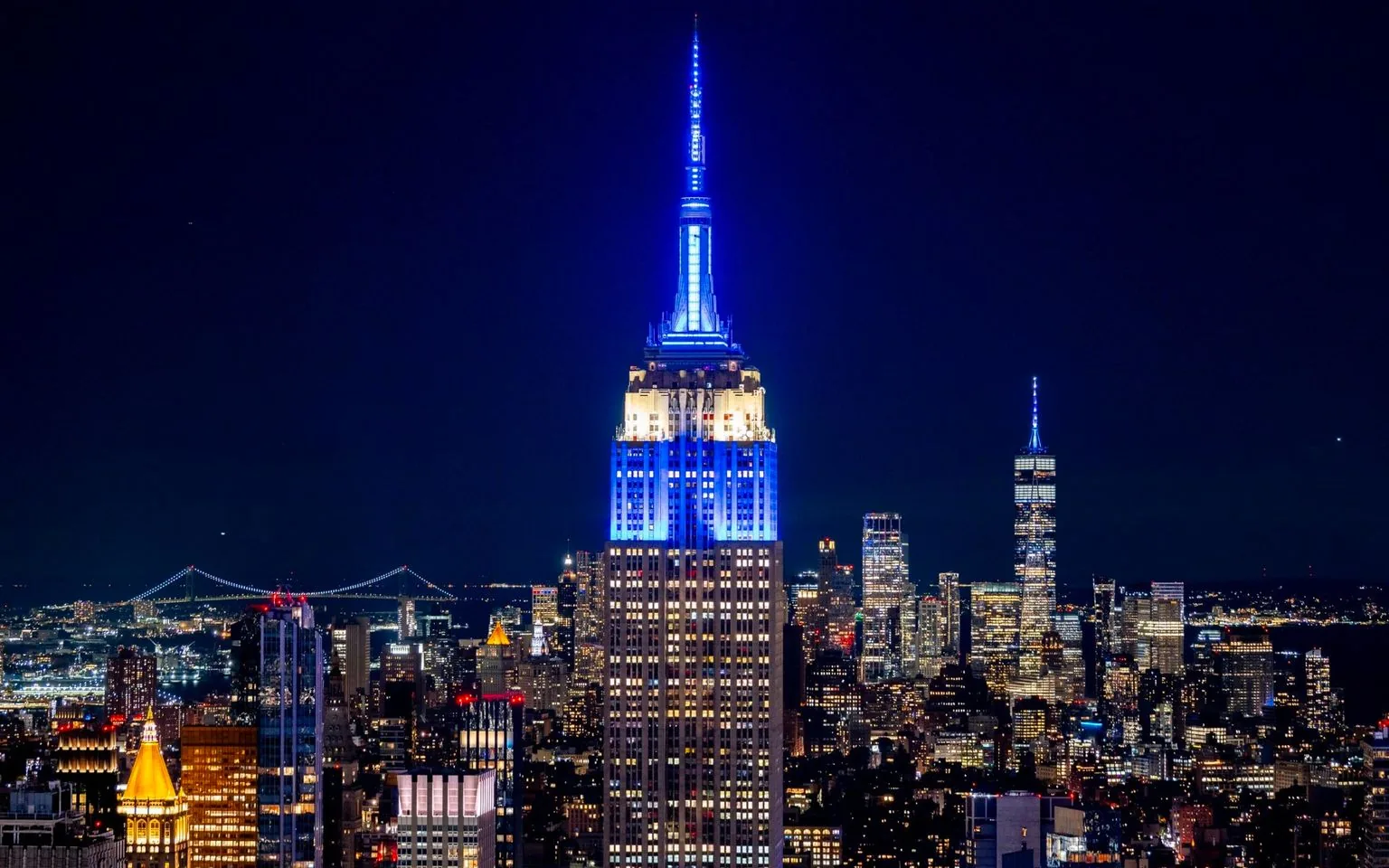The image showcases a breathtaking night view of New York City, with the Empire State Building as the central focus. The building is illuminated in vibrant blue, casting a striking contrast against the dark night sky. Surrounding it are countless other skyscrapers, all aglow with lights, creating a vibrant cityscape that extends into the distance. In the background, the One World Trade Center is visible, also brightly lit and standing tall among the city’s skyline. The composition captures the essence of New York City’s iconic architecture and its bustling, lively atmosphere. The lighting emphasizes the vertical lines of the buildings, highlighting their grandeur. The overall mood of the image is dynamic and electric, reflecting the city’s reputation as the city that never sleeps.