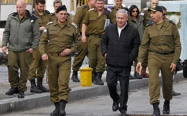 The image shows Benyamin Netanyahu walking alongside a group of military personnel. They are outdoors, possibly at a military base or similar setting. Netanyahu is at the center, wearing a black jacket, dark pants, and a light-colored shirt, exuding a casual and approachable demeanor. He is surrounded by soldiers in uniform, which includes olive green attire and berets, suggesting they are members of the military. The group appears to be engaged in conversation, with some members looking towards Netanyahu, indicating his central role in the interaction.
The background features industrial elements like a yellow bollard and concrete structures, which further emphasize the setting as a base or dock. The mood is professional yet informal, with everyone appearing attentive and engaged. The lighting is natural, likely indicating daytime, and colors in the scene are generally muted, dominated by the earthy tones of the military uniforms and the neutral tones of the surroundings.