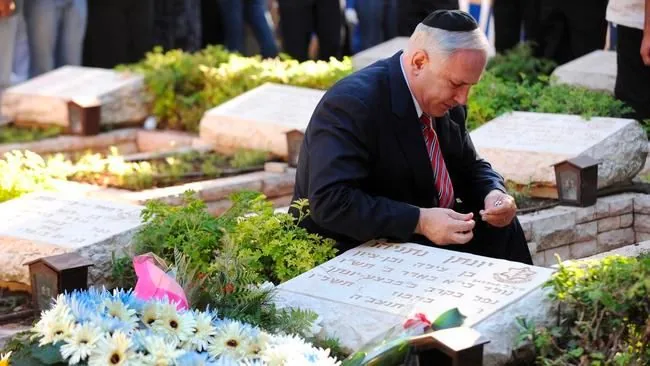 1) Rich description: In the image, Benyamin Netanyahu is seen at a somber moment, kneeling by the grave of his brother, Yoni. The setting is a cemetery, with several gravestones visible in the background. Netanyahu is wearing a dark suit with a red and white striped tie, and a kippah on his head, indicating the reverence of the moment. His expression is reflective and solemn as he appears to be engaged in a moment of personal reflection or prayer. There are flowers placed on Yoni's grave, including a bouquet of white and yellow daisies with pink wrapping. The gravestones are made of a light-colored stone, with Hebrew inscriptions visible on them. The lighting is natural, suggesting the image was taken during the daytime, and the surrounding greenery adds a sense of peace to the scene.
2) On-screen text block:
- Title: "..."