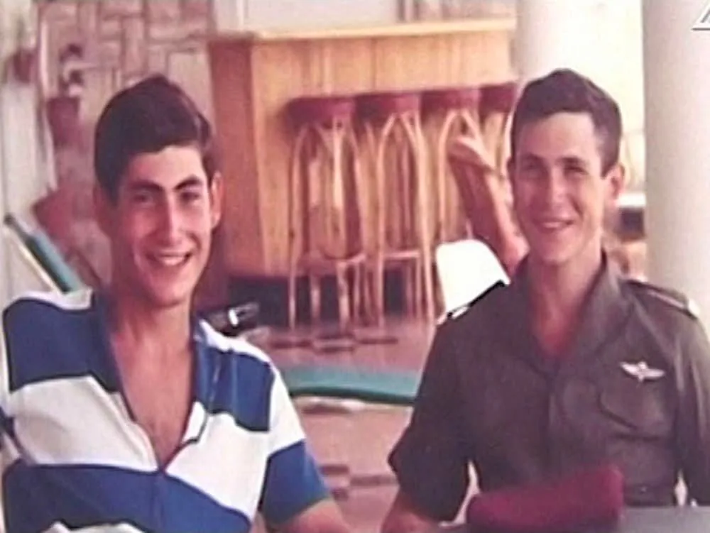 The image features two young men, identified as Benyamin Netanyahu and his brother Yoni. They are seated in a casual indoor setting. Benyamin, on the left, wears a relaxed blue and white striped shirt, exuding a laid-back vibe. He smiles broadly, creating a light and friendly atmosphere. Yoni, on the right, is in military attire, with a uniform that includes a dark green shirt featuring a discernible emblem on the shoulder.
The background includes wooden barstools and a counter, suggesting a recreational or social environment. The lighting is soft and warm, enhancing the vintage feel of the photograph, which appears to be from several decades ago. The brothers convey a sense of camaraderie and familial bond, captured in a moment of leisure.
