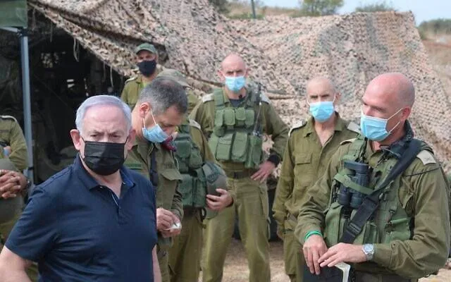 The image shows Benyamin Netanyahu in an outdoor military setting. He is surrounded by several individuals dressed in military uniforms. The group is standing in front of a camouflage netting, suggesting a field or training area. Netanyahu, wearing a black mask and a dark navy polo shirt, appears to be in conversation with the military personnel. The soldiers are wearing masks and tactical vests, with some of them holding notebooks or equipment. The atmosphere seems serious and professional, reflecting a military briefing or inspection. The lighting is natural, indicating that it is likely daytime. The image composition focuses on Netanyahu in the foreground, while the soldiers are positioned around him. The overall tone suggests a strategic or security-related discussion.