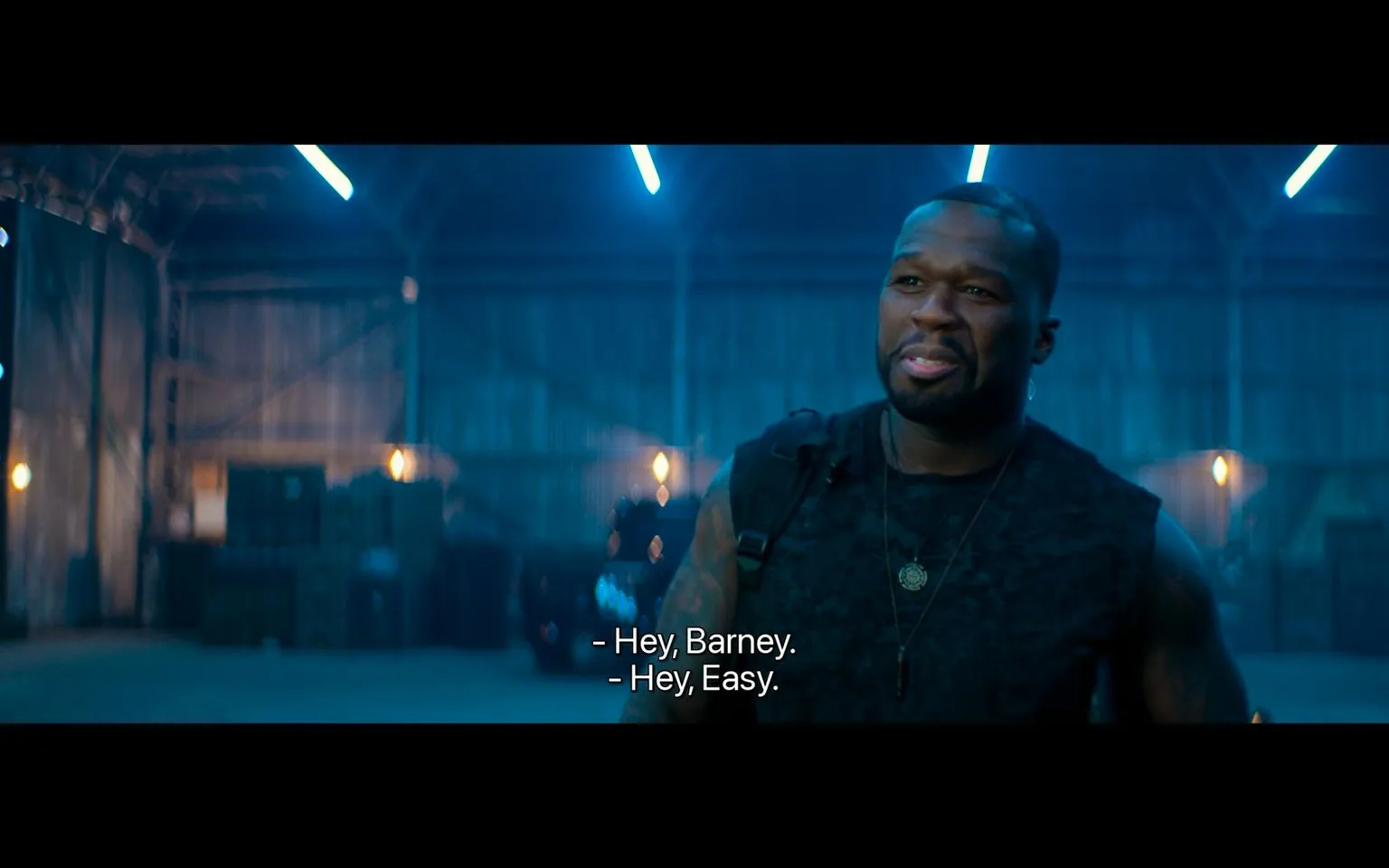 The image shows a scene set within a dimly lit warehouse, where 50 Cent is featured prominently. He is wearing a dark sleeveless shirt, displaying a muscular build. A necklace is visible around his neck, adding a subtle touch to his outfit. The background is filled with crates and subdued lighting, creating a clandestine and intense atmosphere appropriate for an action or negotiation scene. The lighting is predominantly cool with blue tones, contributing to the moody ambiance of the setting.
50 Cent appears relaxed yet focused, engaging in a conversation with someone referred to as "Barney." The interaction is informal, as indicated by the subtitles: "Hey, Barney." "Hey, Easy." This suggests familiarity between the characters. The composition features 50 Cent slightly to the right, allowing for a glimpse of the warehouse interior behind him. The setting, combined with his attire and expression, suggests a moment of casual yet purposeful dialogue, possibly within an action or crime-related narrative.