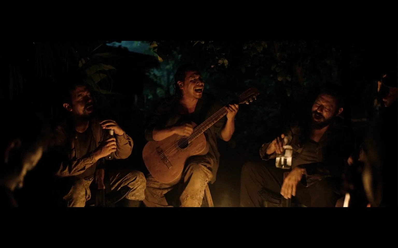 This image captures a nighttime scene in a jungle setting. A group of men are gathered around a campfire, creating an intimate and relaxed atmosphere. Centered in the image, one man is playing a guitar, singing passionately, while others join in with drinks in hand. The soft glow of the firelight casts warm tones on their faces, enhancing the mood of camaraderie and storytelling. Their clothing appears rugged and worn, suggesting they may be explorers or soldiers in a remote area. The background is shrouded in darkness, with hints of dense foliage, adding to the secluded jungle environment. The scene conveys a sense of brotherhood and shared experience, likely a moment of respite amidst challenging circumstances.