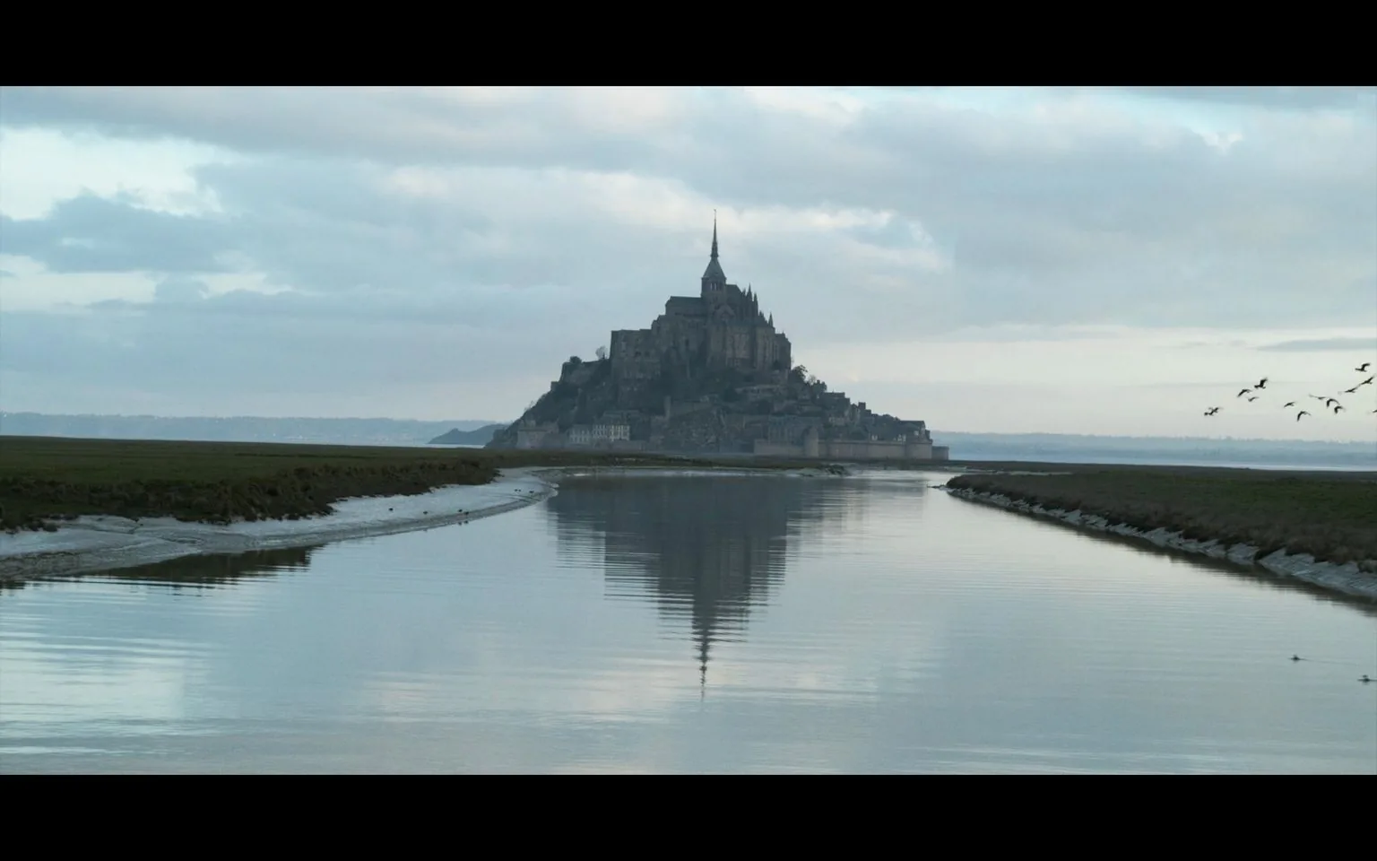 The image showcases a majestic and historic structure, Mont Saint-Michel, located on an island off the coast, with the stunning sky and water creating a serene atmosphere. Rising dramatically from the landscape, the island is dominated by its iconic abbey, which features intricate Gothic spires and stonework, creating a picturesque silhouette against the clouds. The reflection of the structure in the calm water adds depth and symmetry to the composition. The color palette is subdued, with soft blues and grays, enhancing the tranquil mood. Birds can be seen flying on the right side, adding a touch of natural life to the scene. The surrounding landscape is flat and grassy, leading the eye towards the towering architectural marvel. The overall scene is peaceful, capturing the timeless beauty of this UNESCO World Heritage site.
- Captions / subtitles: "Hi Oliver!!!", "You guys can come over for my inauguration", "Elton John's doing Circle of Life", "Taylor Swift is singing Star Spangled Banner"