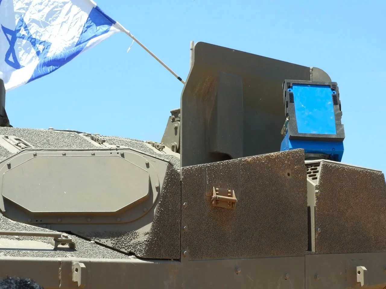This image displays a close-up of a military vehicle equipped with the Trophy defense system. The vehicle is covered in a rugged, textured armor that provides protection and durability. A prominent feature is the bright blue component of the Trophy system, mounted on top of the vehicle. This system is designed to intercept and neutralize incoming threats, enhancing the vehicle's defense capabilities.
An Israeli flag is visible, suggesting the vehicle's origin or affiliation with the Israeli Defense Forces. The scene is set outdoors under a clear blue sky, indicating daytime conditions. The overall appearance is utilitarian, with the vehicle's surface featuring various angular panels and hatches. The robust design and strategic placement of the defense system components highlight the vehicle's purpose for military operations.