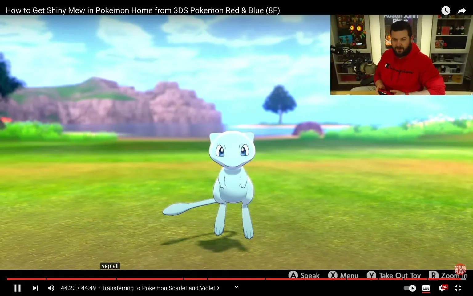 The image is a frame from a YouTube video titled "How to Get Shiny Mew in Pokemon Home from 3DS Pokemon Red & Blue (8F)" on the channel "Austin John Plays." In the main part of the image, a Shiny Mew is prominently displayed against a colorful outdoor setting within a video game environment, featuring grassy plains and distant hills under a clear blue sky. The Mew is light blue, indicating its shiny variant, and is floating cheerfully in the center of the frame.
In the top right corner, there is a small inset of a man, presumably Austin John, sitting in a room with shelves of collectibles and gaming paraphernalia in the background. He is wearing a red Champion hoodie and appears to be holding a game controller. The room is well-lit and has a casual, gamer-friendly atmosphere.
On-screen text includes button prompts like "A Speak," "X Menu," "Y Take Out Toy," and "R Zoom In," typical for a game interface. The video is near completion, with a timestamp of "44:20 / 44:49," and the video description notes, "Transferring to Pokemon Scarlet and Violet." Subtitles display the phrase "yep all," suggesting a response to an in-game event or action. The video seems to be a tutorial or guide focused on transferring a Shiny Mew within Pokémon games.