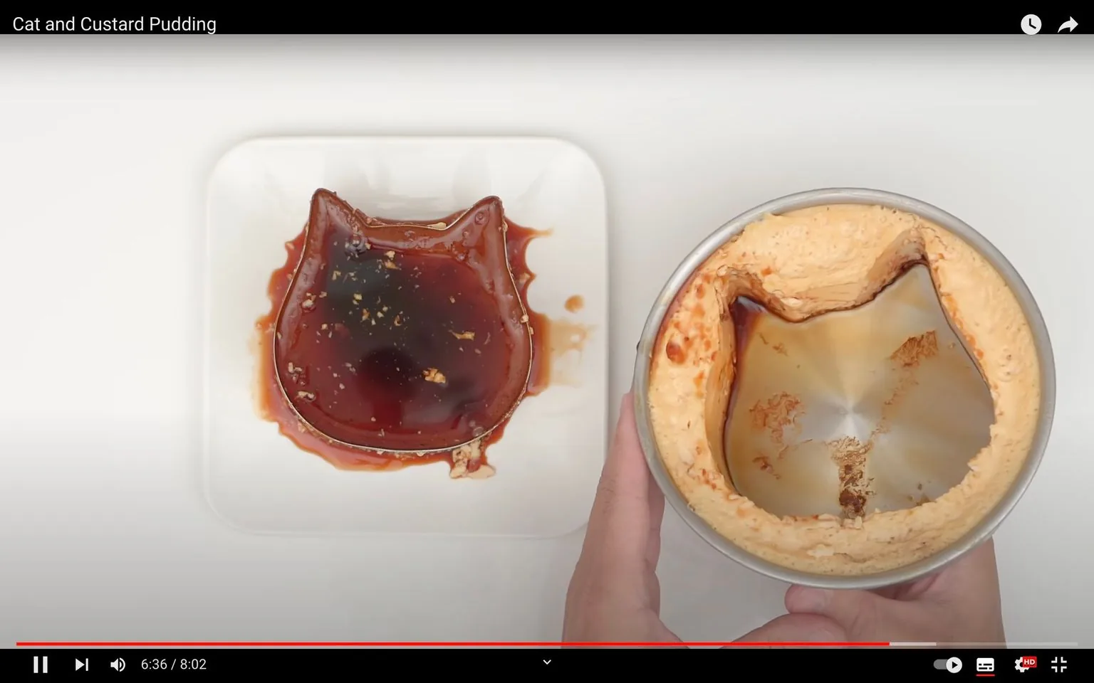 The image shows a still from a video titled "Cat and Custard Pudding." It features a custard pudding that has been molded in the shape of a cat's face. The pudding is placed on a white square plate, with a rich, dark caramel sauce drizzled over its surface, giving it a shiny and appetizing appearance. To the right, there is an emptied mold, indicating the pudding was recently removed from it. The mold also has the cat shape, with remnants of custard and caramel sauce visible inside. The setting appears to be a kitchen or a food preparation area, with a clean, white background that accentuates the pudding's colors. The scene is brightly lit, focusing on the dessert's texture and form, suggesting a tutorial or cooking demonstration context.