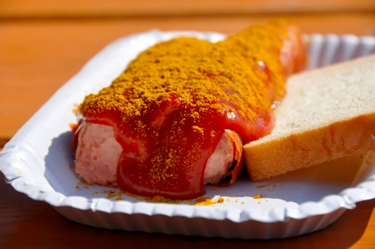The image displays a serving of currywurst, a popular German dish. The dish consists of a sausage covered in a generous amount of ketchup or a similar red sauce, topped with a layer of curry powder. It is served on a small, white, disposable paper plate. Next to the sausage is a slice of white bread, possibly a side accompaniment to the main dish. The setting appears to be outdoors, as suggested by the bright, natural lighting that highlights the textures and colors of the food. The background is slightly blurred, drawing focus to the vivid contrast between the red sauce and the yellow curry powder. The overall presentation is casual, typical of a street food or fast food setting.