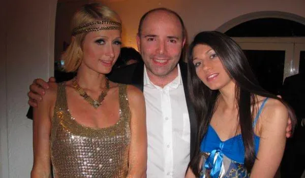 Paris Hilton, Pierre Woodman, and another woman are pictured together at an indoor event. Paris Hilton is on the left, wearing a glittering gold dress with sequins and a matching headband. Her hair is styled in an elegant updo, and she accessorizes with a bold necklace. In the center is Pierre Woodman, dressed in a classic black suit with a white shirt, smiling warmly at the camera. The woman on the right is wearing a vibrant blue dress with a patterned design and has long, straight dark hair. The setting appears to be a lively evening gathering, with a warm, ambient light enhancing the festive atmosphere. The background shows a simple, arched doorway, indicative of an indoor venue, possibly a private party or event. The overall mood is celebratory and friendly, with everyone posing closely together, suggesting familiarity and camaraderie.