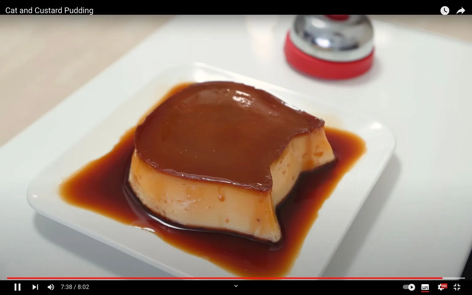 The image is a video frame showcasing a delectable dessert prominently placed on a white square plate. The dessert appears to be a custard pudding with a rich caramel sauce glistening on top. The pudding has a smooth, creamy texture, and a portion has been taken out, revealing its soft interior. Surrounding the custard is a generous pool of caramel sauce, adding an indulgent touch to the presentation. In the background, there is a small red and silver item, possibly a bell or another culinary tool, slightly out of focus. The scene is set in a brightly lit area, enhancing the dessert's inviting appearance. The title text at the top reads "Cat and Custard Pudding," suggesting the theme or recipe featured in the video.