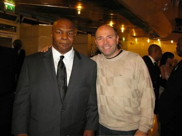 Mike Tyson and Pierre Woodman are posing together in an indoor, formal setting. Mike Tyson is wearing a dark suit with a white shirt and a black tie, exuding a formal and composed demeanor. Pierre Woodman stands beside him wearing a light beige sweater with a subtle pattern and a collared shirt underneath. The setting suggests a social or professional event, as indicated by the elegant attire and the presence of other people in the background, who appear to be engaged in conversations or mingling. The lighting is warm and ambient, casting a comfortable glow over the scene. The atmosphere appears cordial and relaxed, with both individuals smiling slightly, suggesting a friendly interaction. The background displays some architectural elements and lighting fixtures, adding to the sophisticated ambiance of the event.