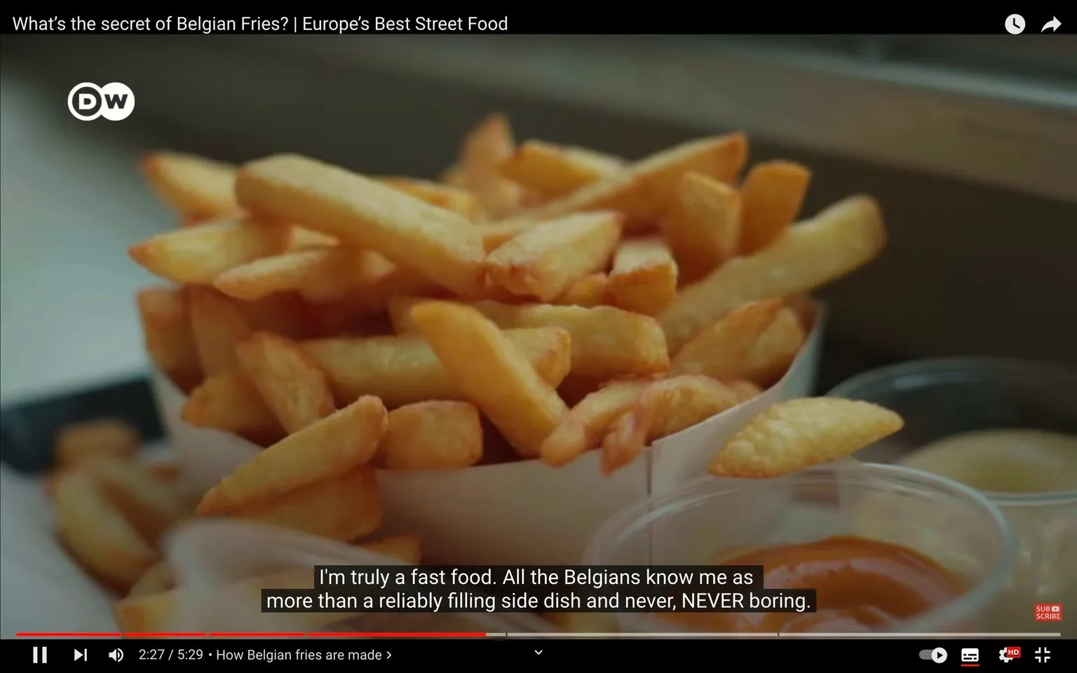 The video frame features a close-up of a serving of Belgian fries, which are showcased in a paper cone. These fries appear golden and crispy, characteristic of traditional Belgian preparation. Accompanying the fries are small cups filled with various sauces, suggesting a selection for dipping. The lighting in the scene is natural, highlighting the texture and color of the fries, making them look appetizing. The composition centers on the fries, drawing attention to their quality and appeal as a popular street food.
The video, titled "What's the secret of Belgian Fries? | Europe's Best Street Food," is from the DW channel on YouTube. The subtitles convey a narrative that emphasizes Belgian fries as a beloved and never boring fast food, highlighting their significance beyond just being a filling side dish. The context suggests an exploration into what makes Belgian fries special, likely discussing their preparation and cultural importance.