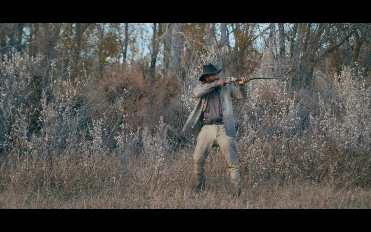 The image features a person standing in a forest with a rifle aimed forward, suggesting a moment of action or potential conflict. The person is dressed in attire typical of a Western setting, with a wide-brimmed hat, long coat, and rugged pants, reinforcing a historical or thematic Western ambiance. The coat is open, revealing a dark shirt underneath, and the person stands with a wide stance, indicating readiness and stability. The scene is set in a dry, sparse forest with tall grass and barren trees, implying an autumn or early winter season due to the lack of foliage. The colors in the image are muted, with shades of brown and gray dominating, enhancing the rustic and gritty feel of the setting. The lighting is natural, likely indicating late afternoon or early evening as shadows are present but not harsh. There is no on-screen text or branding visible, allowing the scene to focus entirely on the action unfolding.