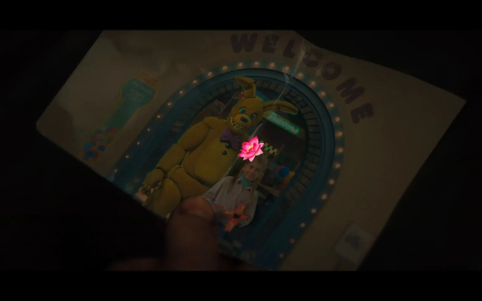 The image depicts a photograph held by a hand, showcasing a scene from "Five Nights at Freddy's." In the photo, a child is standing next to an animatronic character, which resembles a large, yellow rabbit with a bow tie, set against a backdrop of a festive archway adorned with lights. Above the archway, the word "WELCOME" is prominently displayed. The child is seen holding a toy, and both appear to be in a playful setting possibly resembling a themed pizzeria, which is a common setting in the "Five Nights at Freddy's" series. The lighting is dim, with a focus on the photograph, giving it a nostalgic or mysterious atmosphere. The overall composition suggests a blend of innocence and the unsettling nature typical of the franchise's aesthetic.