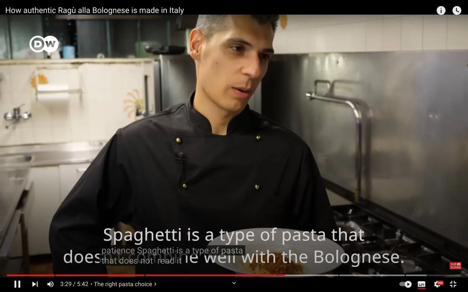 The video frame is from a YouTube video titled "How authentic Ragù alla Bolognese is made in Italy," published by DW. It features an indoor kitchen setting where a chef is being interviewed or explaining a topic. The chef is wearing a black chef's jacket with gold buttons and is looking slightly to the side while speaking. He holds a plate in one hand, which possibly contains pasta, aligning with the video's theme.
The background shows typical kitchen equipment, like a metal backsplash and a sink, suggesting a professional kitchen environment. The lighting is bright, focusing on the chef, who appears engaged and serious in his explanation. The subtitles on the video state, "Spaghetti is a type of pasta that does not read it well with the Bolognese," indicating a discussion about pasta types suitable for Ragù alla Bolognese.
The video is formatted with standard YouTube interface elements, including a progress bar at the bottom, indicating the video is 3 minutes and 29 seconds into a total duration of 5 minutes and 42 seconds. The DW logo is visible on the top left corner, reinforcing the channel's branding.