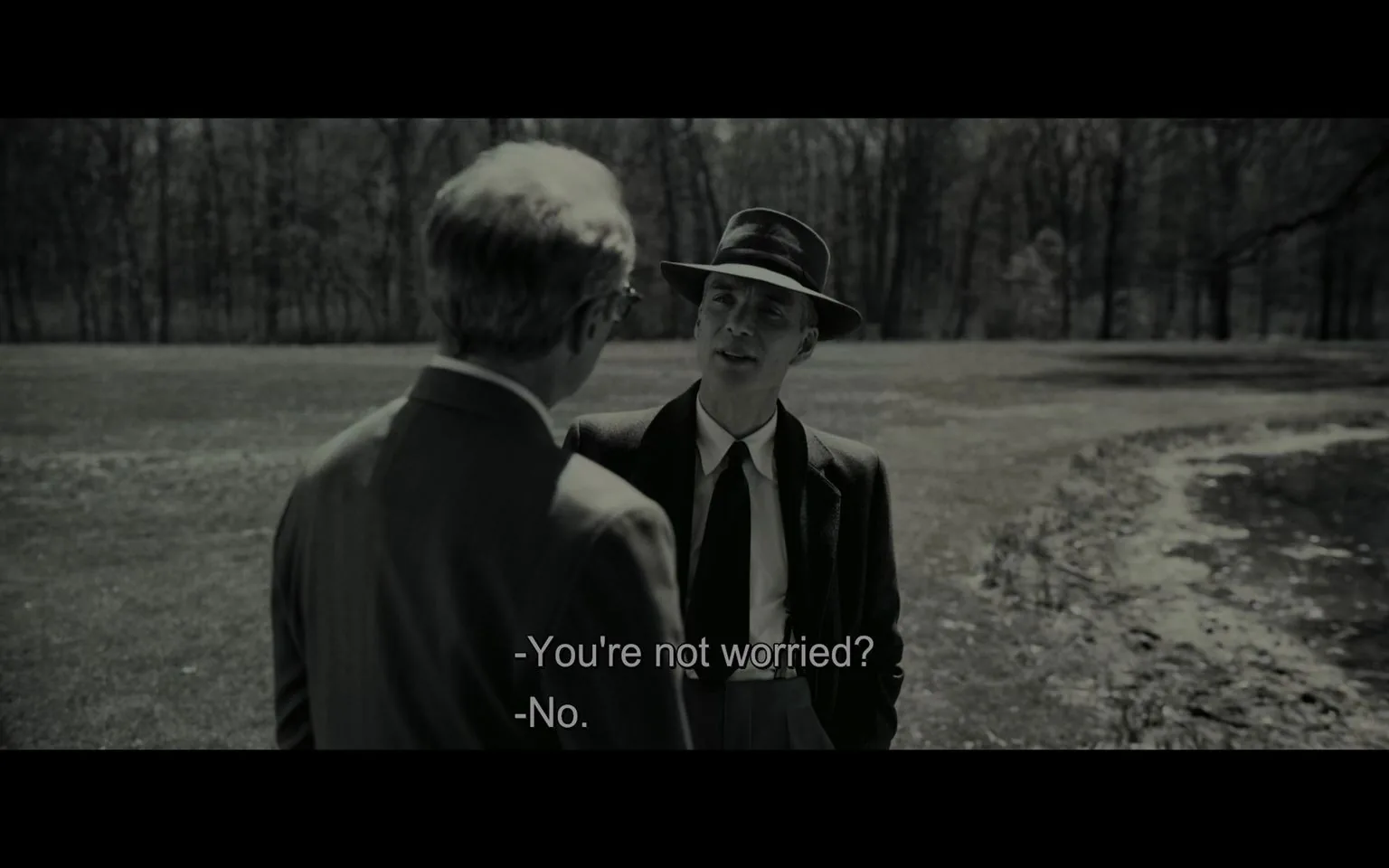 In this black-and-white image, Cillian Murphy is portrayed as Oppenheimer, engaged in conversation with another man. The scene is set outdoors, possibly in a park or open field, with trees in the background adding to the serene yet tense atmosphere. Cillian Murphy, dressed in a classic suit and tie, also wears a fedora, giving him a distinct mid-20th-century appearance. He stands with one hand in his pocket, displaying a calm demeanor as he responds to the other man's concern. The other man, seen from the back, is also in formal attire, suggesting a professional or serious discussion. The lighting is natural, emphasizing the shadows and contrasts typical of black-and-white cinematography. The subtitles reveal part of their dialogue: "-You're not worried?" "-No." This exchange suggests a conversation about a potentially serious matter, with Murphy's character displaying confidence or nonchalance. The overall composition and attire reflect the historical setting of the film "Oppenheimer."
- Captions / subtitles: "-You're not worried?" "-No."