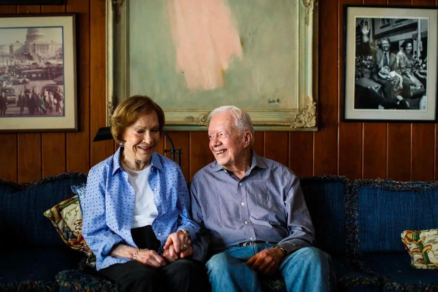 Jimmy Carter is sitting beside his wife, both exuding warmth and affection in a cozy living room setting. The couple is seated on a dark blue couch with patterned cushions, creating a comfortable and intimate atmosphere. Jimmy Carter is wearing a striped shirt with light blue jeans, his expression is joyful as he looks lovingly at his wife. She is wearing a light blue polka dot shirt over a white top, smiling warmly. The background features wooden paneled walls adorned with framed photographs—a color image of a busy street scene and a black-and-white photograph of a couple waving from a car. A muted abstract painting hangs prominently in the center. The lighting is soft and natural, highlighting the gentle and serene mood of the scene. The overall composition conveys a sense of enduring love and companionship.