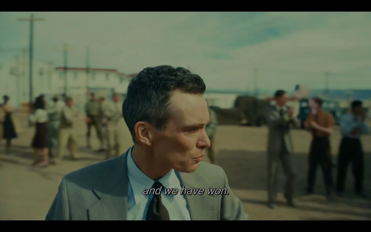 In this video frame, Cillian Murphy is shown portraying J. Robert Oppenheimer. He is standing outdoors in a setting reminiscent of a mid-20th-century military or scientific complex. The focus is on Murphy, who is dressed in a mid-grey suit with a white shirt and a dark tie, exuding a formal and serious demeanor. His expression is determined, capturing a moment of significance. The background features several people in period-appropriate attire, including military uniforms, suggesting a scene of celebration or announcement. Some individuals appear to be clapping, and an American flag is subtly visible among the crowd. The lighting is natural, suggesting daylight, which complements the historical ambiance of the scene. The subtitle at the bottom of the frame reads, "and we have won," indicating a victorious or triumphant moment. This frame is from the film "Oppenheimer."
- Captions / subtitles: "and we have won."