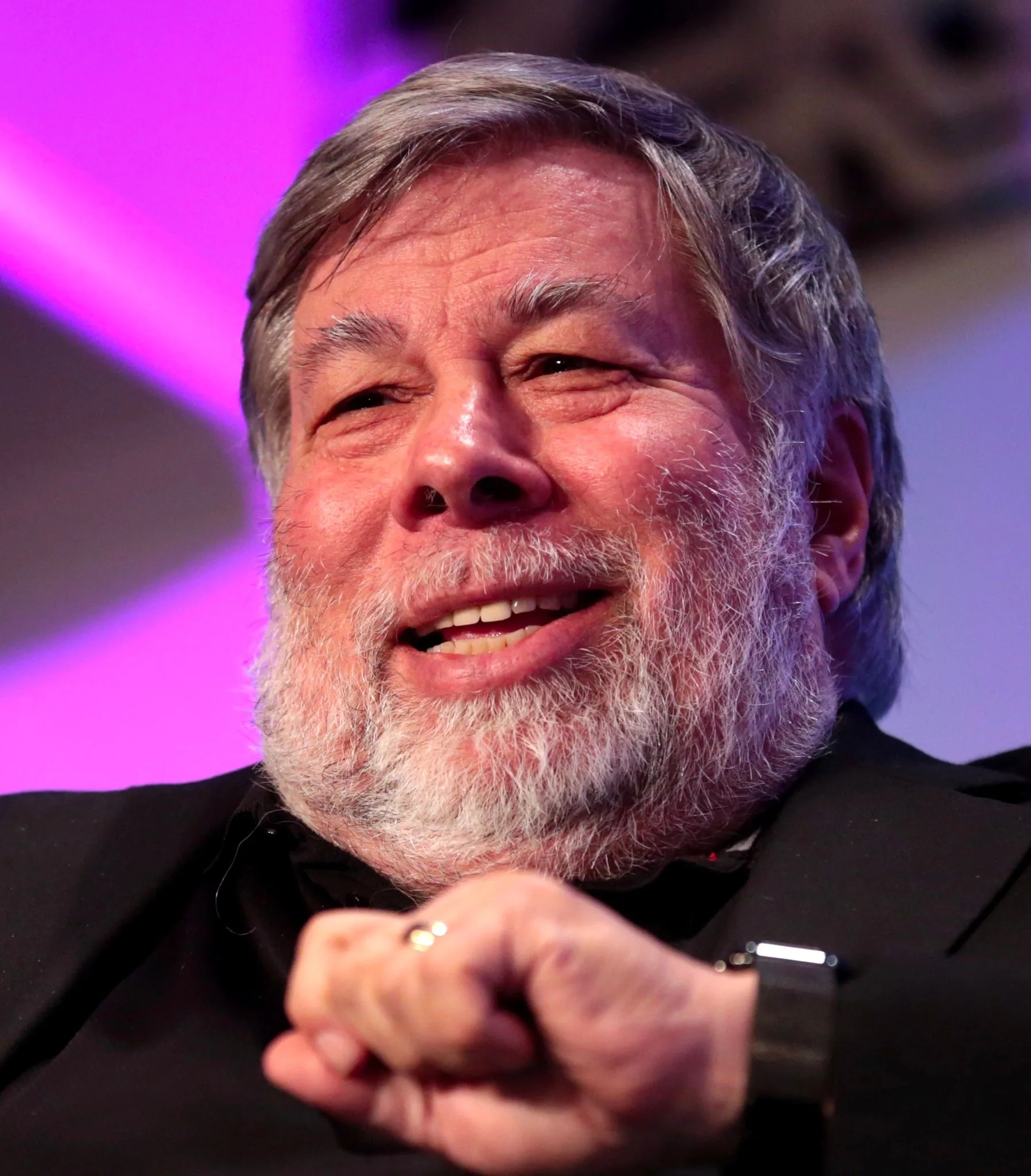 The image shows an older man with a white beard and gray hair, smiling broadly. He appears to be engaged in a conversation or an interview, suggested by his expression and the visible gesture of his hand, which is partially clenched. The lighting in the image is warm, casting a pleasant glow on his face, and the background is softly blurred with a purple hue, indicating an indoor setting, likely at an event or conference. He is wearing a dark colored shirt or jacket, and a watch is visible on his wrist. The atmosphere of the image is lively and positive, with the man looking relaxed and approachable. There is no visible text or branding in the image.