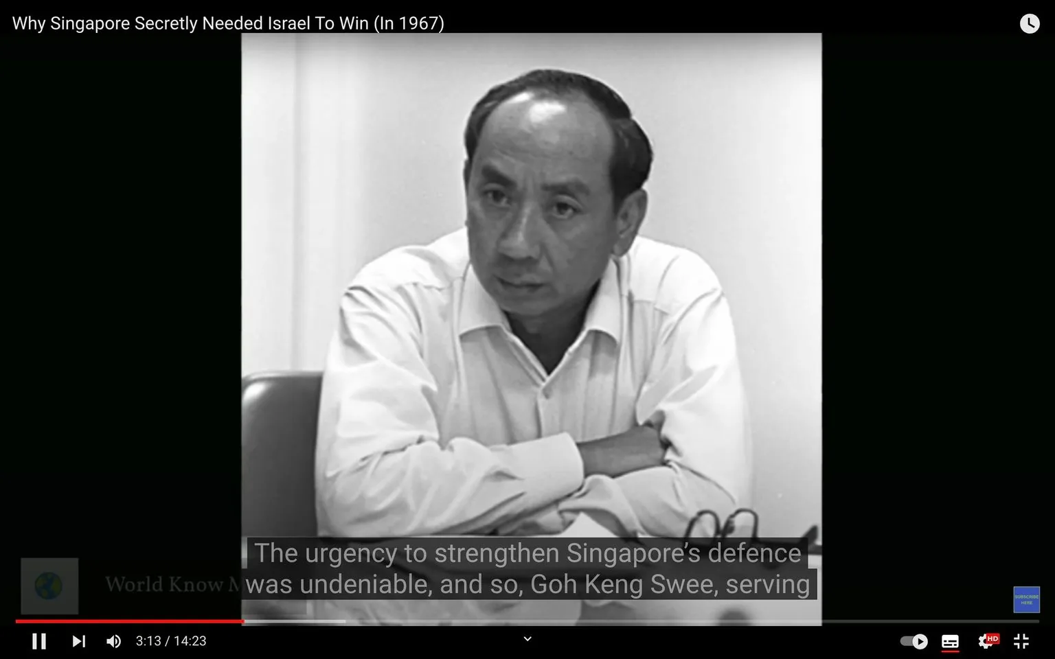 The image is a black and white video frame from a YouTube video titled "Why Singapore Secretly Needed Israel To Win (In 1967)." It features a man identified in the context as Goh Keng Swee, who appears to be in an indoor setting, likely an interview or meeting. He is seen sitting at a table, wearing a light-colored, long-sleeved shirt with his arms crossed on the table. His expression is serious, suggesting engagement or deep thought in the discussion. The black and white tone of the image adds a historical or documentary feel, aligning with the video's topic about Singapore's defense strategies in 1967. Subtitles at the bottom of the frame read, "The urgency to strengthen Singapore’s defence was undeniable, and so, Goh Keng Swee, serving," indicating a focus on defense matters. The setting is simple, with a plain background, emphasizing the subject's presence and the gravity of the discussion.