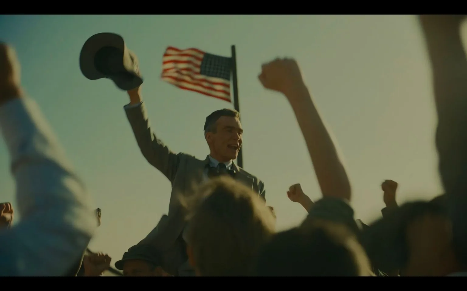 Cillian Murphy is depicted in this image from the film "Oppenheimer." He is surrounded by an enthusiastic crowd, raising their fists in celebration. Murphy, dressed in a suit and tie, holds a hat in one hand and is smiling broadly, conveying a sense of triumph or achievement. The scene is set outdoors, with an American flag waving prominently in the background, suggesting a patriotic or historical context. The lighting is warm and natural, giving the scene a vibrant and uplifting atmosphere. The composition focuses on Murphy, elevated above the crowd, highlighting his central role in the moment. The overall mood is jubilant, capturing a significant, victorious moment.