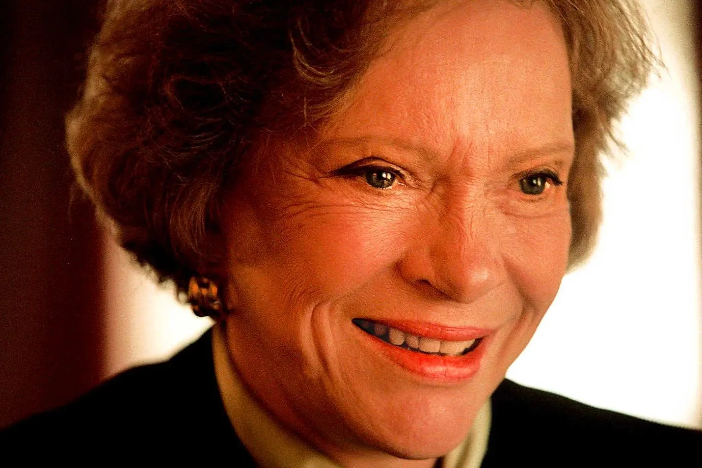 The image is a close-up portrait of a person with a warm and friendly expression, captured indoors. The lighting is soft and highlights the subject's facial features, creating a welcoming and inviting atmosphere. The individual has short, wavy hair and is wearing a black outfit with subtle jewelry, including earrings that add a touch of elegance. The background is softly blurred, which keeps the focus on the person's face. The overall composition is balanced, emphasizing the genuine smile and the warmth of the eyes. The colors are rich and natural, enhancing the subject's complexion and the ambient light in the room. The mood conveyed is one of kindness and approachability.