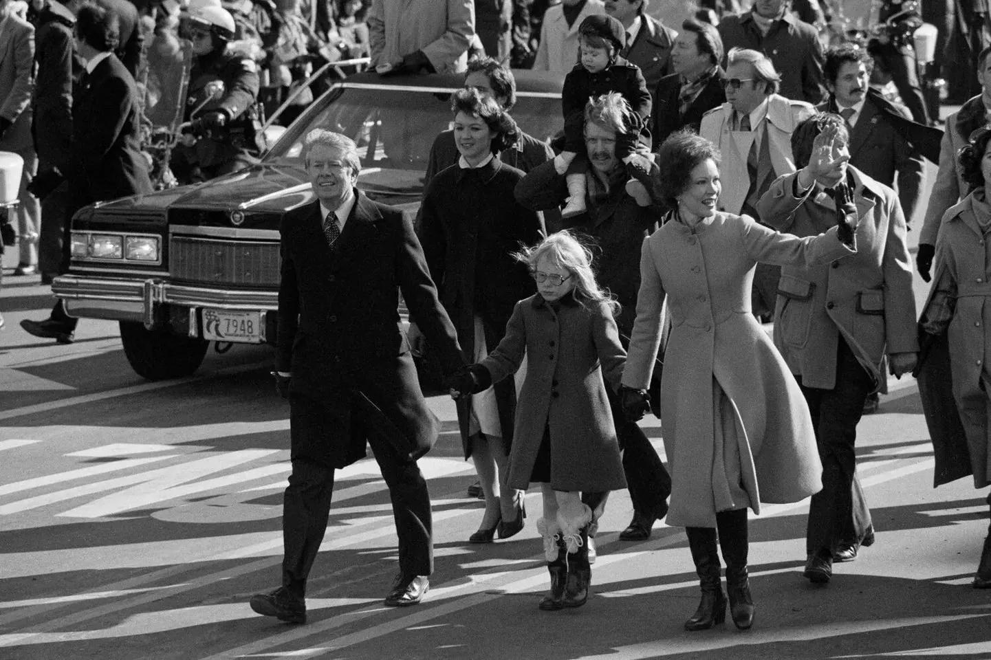 In this historical black-and-white photograph, Jimmy Carter is seen in a celebratory parade, walking with a group of people, likely during his inauguration as President of the United States. Jimmy Carter is in the foreground, smiling as he strides confidently down the street, holding hands with others, suggesting a family or close group moment. Beside him, a woman in a long coat waves to the crowd, and a young girl, possibly a family member, walks alongside them. The atmosphere is lively, with numerous people in the background, some of whom appear to be part of a procession or parade. There is a visible police presence, with officers on motorcycles in the background, contributing to the event's security measures. The scene is bustling with energy, as bystanders and participants soak in the celebratory mood. The clothing styles are reflective of the era, with long coats and formal attire being prominent. Behind the group, a car follows, emphasizing the importance and official nature of the event.