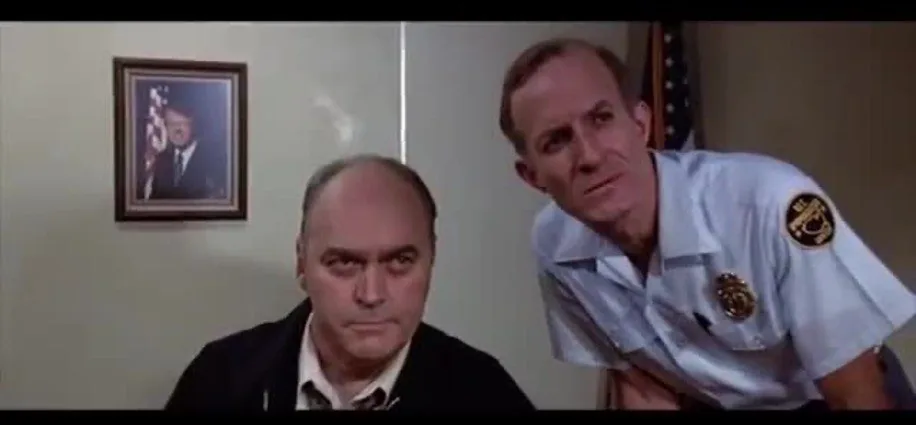 In this indoor scene, two men are present in what appears to be an office setting. The man on the left is dressed in a dark jacket over a white shirt, conveying a serious and focused demeanor. The man on the right is wearing a light blue uniform, possibly indicating a role in security or law enforcement, as suggested by the badge and emblem on his shirt.
In the background, there is a framed photograph on the wall depicting Jimmy Carter, which helps set the political or governmental context of the scene. The American flag is partially visible, reinforcing the official and possibly governmental nature of the location. The lighting is bright and evenly distributed, typical of an office environment. The overall mood is one of concentration or deliberation, as both men appear to be engaged in a conversation or listening intently.