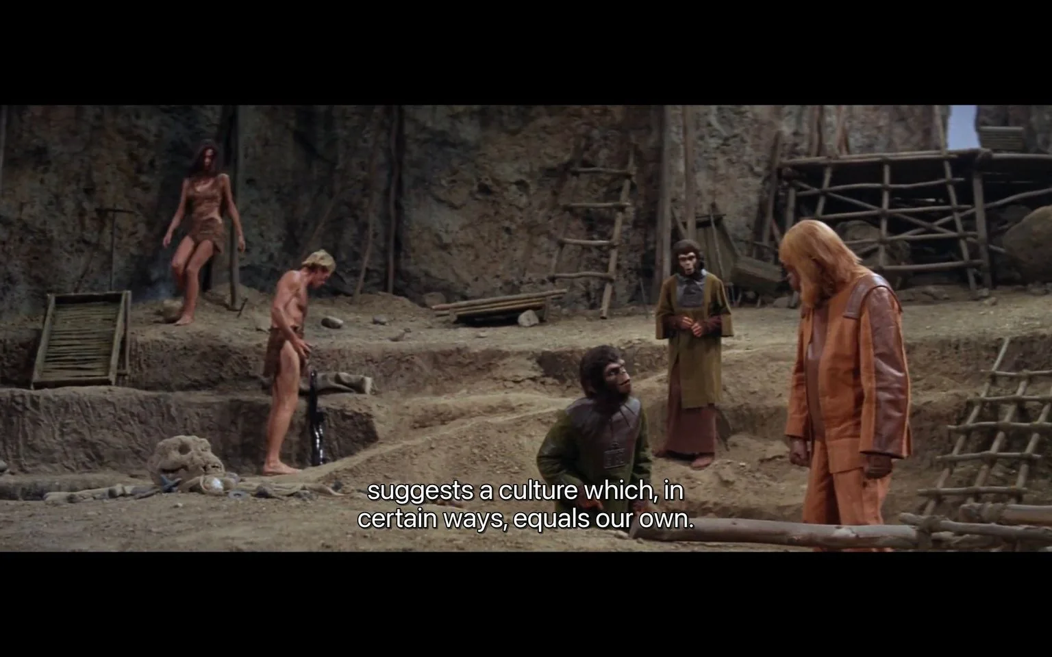The image is a frame from the film "Planet of the Apes." It depicts a scene set in a rustic, outdoor environment, likely an archaeological dig site. Several characters are present, including humans dressed in primitive clothing and apes in more sophisticated attire. The apes are standing and interacting with the humans, suggesting a power dynamic or cultural exchange. The background features a rocky terrain with wooden structures like ladders and platforms. The lighting is natural, highlighting the earthy tones of the surroundings.
One ape, dressed in an orange and brown outfit, stands in the foreground conversing with another ape, who is lower and looking upward. A human male, also in primitive garb, is interacting with the environment to the left. The mood of the scene reflects a mix of curiosity and tension, as suggested by the subtitle: "suggests a culture which, in certain ways, equals our own." This indicates a thematic exploration of cultural parallels between the apes and humans.