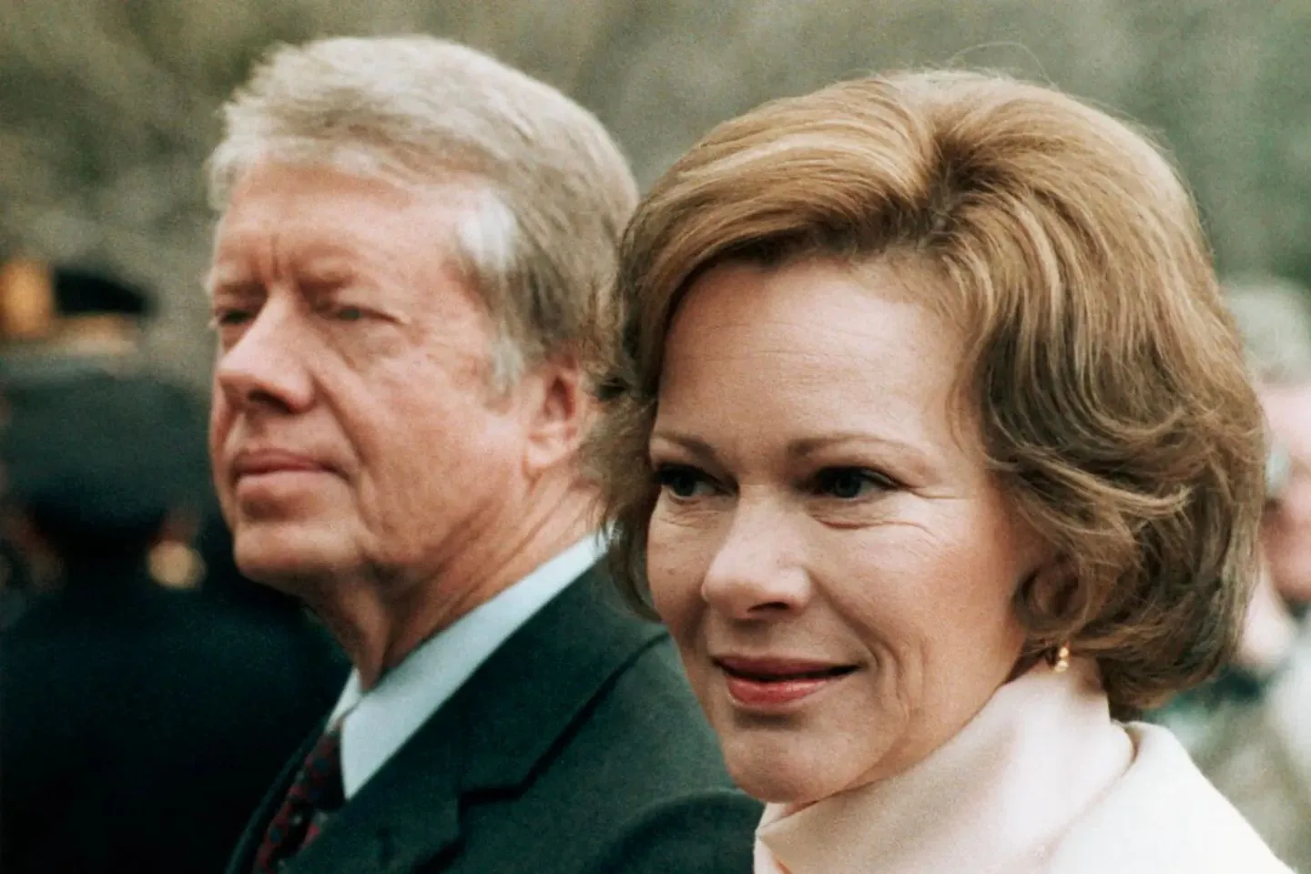 In the image, Jimmy Carter is present alongside a woman outdoors during the day. Jimmy Carter is slightly out of focus in the background, dressed in a formal suit and tie. His expression appears contemplative as he gazes ahead. The woman in the foreground has neatly styled light brown hair and is wearing a light-colored turtleneck with a soft smile on her face, suggesting a pleasant or formal occasion. The background is blurred, but it appears to be a public or ceremonial setting, possibly involving an event with other attendees. The lighting is natural, indicating a daytime setting, and the overall mood suggests a sense of dignity and composure. The image captures a moment of quiet confidence, characteristic of a formal public appearance.