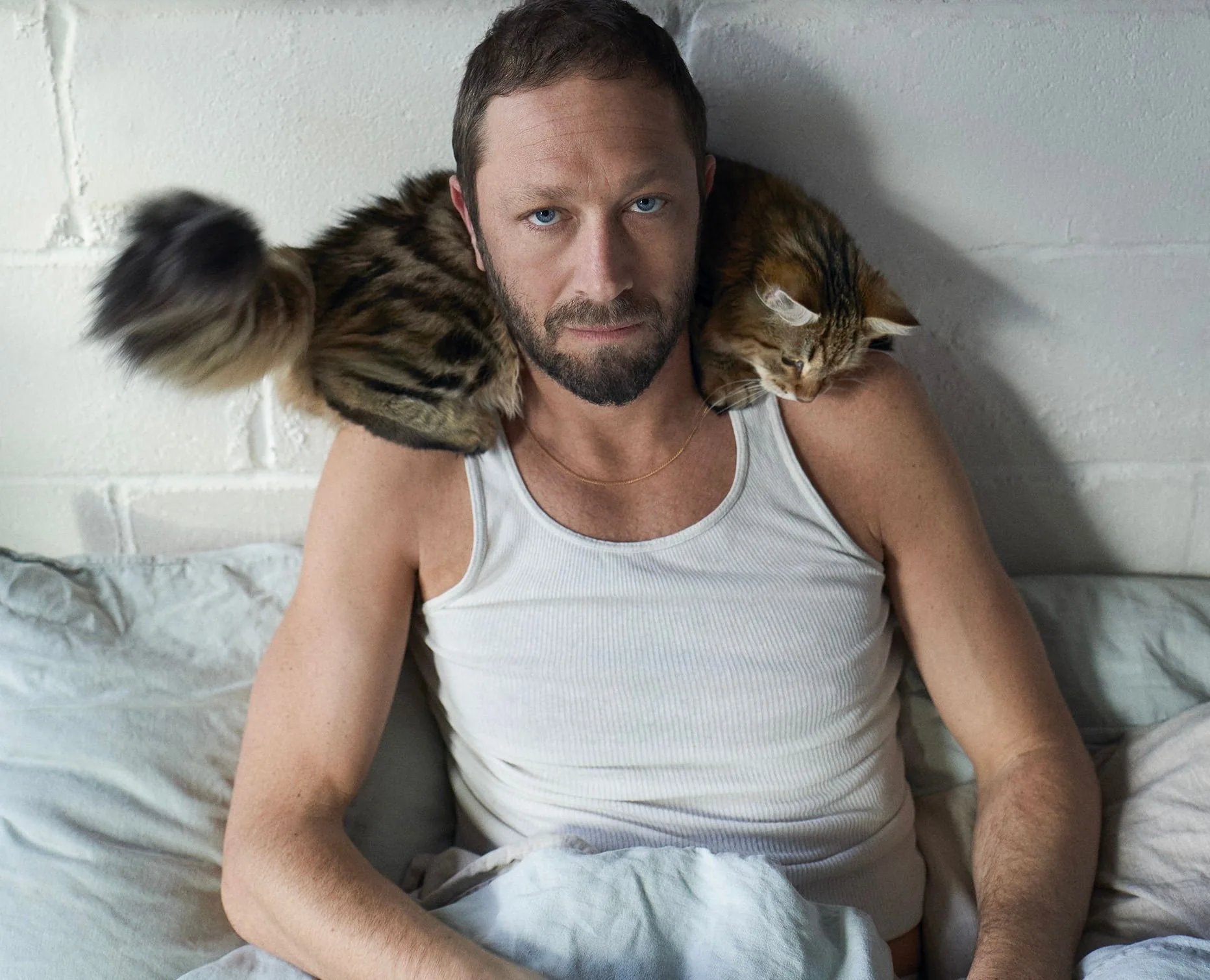 The image features a man sitting up in bed, leaning against a white brick wall. He is wearing a white tank top and appears relaxed, with a calm, thoughtful expression. Resting on his shoulders is a fluffy striped cat, which adds a cozy and intimate feel to the scene. The cat is perched comfortably, with its tail slightly blurred, possibly indicating movement.
The lighting is soft and natural, highlighting the textures of the fabric and the man's features. The setting suggests an early morning or relaxed moment, with the bed covered in light-colored sheets and pillows. The overall mood is serene and personal, conveying a sense of tranquility and companionship between the man and the cat. There are no visible brandings or text in the image.