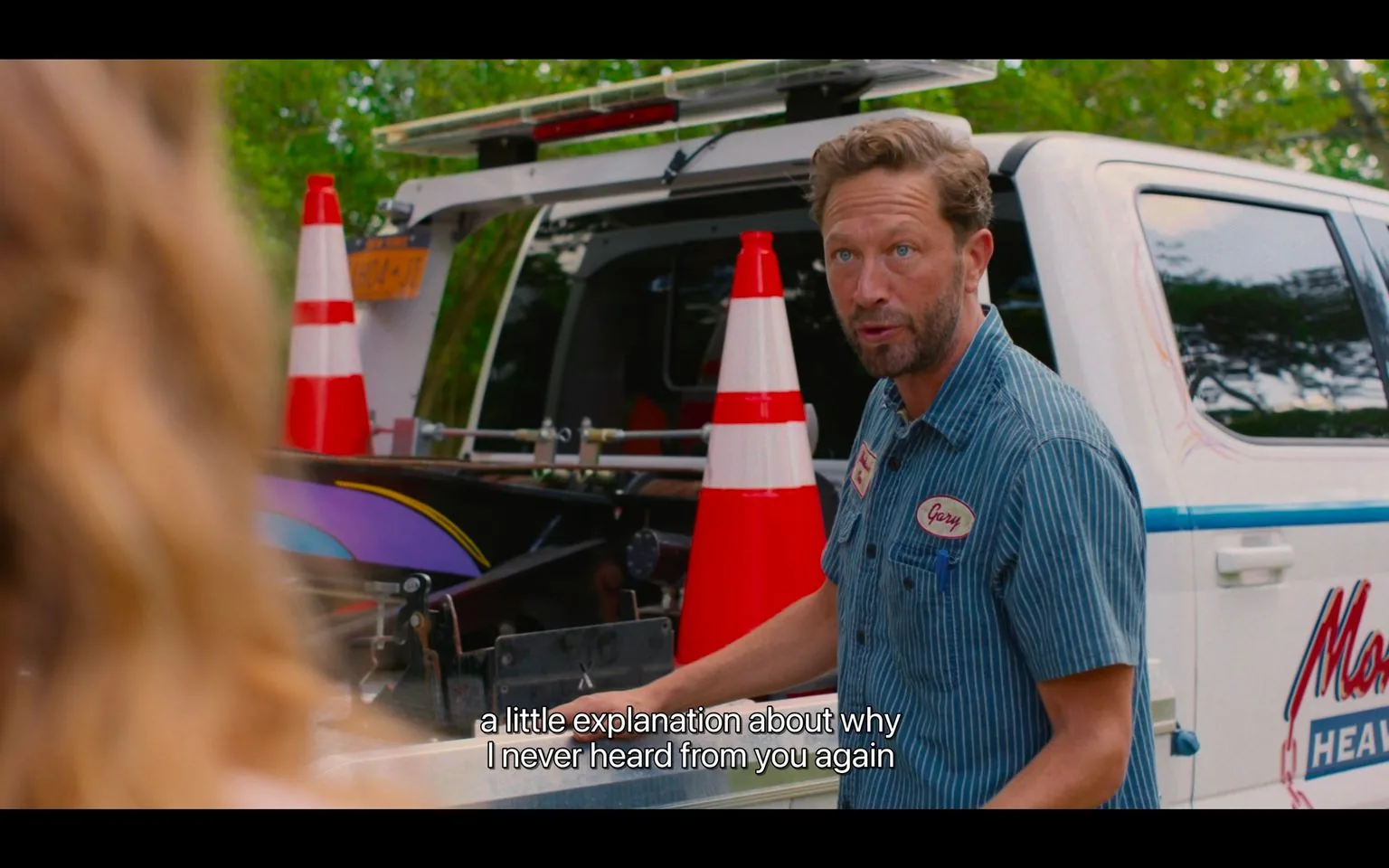 A man with light hair and a short beard, wearing a blue-striped work shirt with a name tag that reads "Gary," is leaning slightly forward, engaging in what appears to be a conversation with another person. His expression is focused and slightly questioning. In the background, a white truck equipped with traffic cones and a visible company logo is parked. The scene is set outdoors with greenery visible in the background, indicating it might be a pleasant day. The truck’s logo includes the word “Heaven,” which hints at a possible business name. The lighting is natural, suggesting it's filmed during the day. The scene captures a moment of dialogue, as evidenced by the subtitle at the bottom: "a little explanation about why I never heard from you again." The overall mood suggests a scene of inquiry or confrontation.
