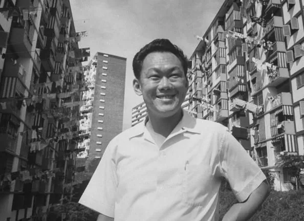 1) Rich description: The image features Lee Kuan Yew, an influential figure in Singapore's history, standing outdoors against a backdrop of high-rise residential buildings. The scene is set during the daytime with clear skies overhead, suggesting a bright and positive ambiance. Lee Kuan Yew is wearing a short-sleeved, light-colored shirt, exuding a casual yet authoritative presence. He is smiling warmly, which conveys a sense of optimism and confidence. The buildings around him are densely packed, characteristic of Singapore's urban landscape, with laundry hanging from the windows, adding a touch of everyday life to the scene. The photograph is black and white, giving it a timeless quality. The composition is balanced, with Lee Kuan Yew centrally positioned, highlighting his importance within the frame and the context of Singapore's development.
2) On-screen text block:
- Title: ""