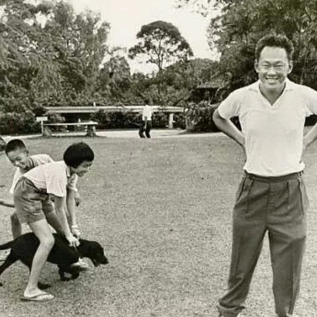 In this black-and-white photograph, Lee Kuan Yew is seen in a relaxed and cheerful mood, suggesting a personal or family outing. He stands at the forefront, wearing a light-colored short-sleeved shirt and dark trousers, with a broad smile and his hands on his hips, conveying a sense of ease and happiness. In the background, a couple of children are playing with a black dog, adding a playful and familial atmosphere to the scene. The setting appears to be a park or garden, with trees and open grass providing a serene backdrop. The overall tone of the image is casual and candid, capturing a moment of leisure. The composition and style reflect a classic, timeless quality typical of mid-20th-century photography. There is no on-screen text visible in the image.