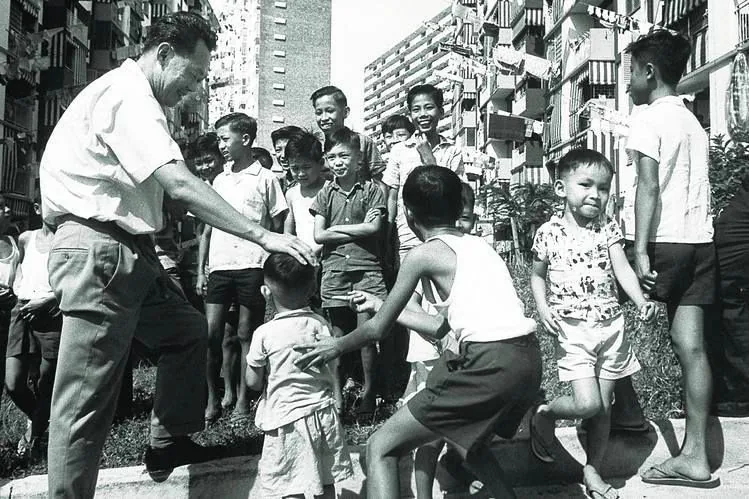 In this black-and-white photograph, Lee Kuan Yew is interacting with a group of children in an outdoor urban setting. Lee Kuan Yew, dressed in a short-sleeved shirt and dark trousers, is seen leaning forward, gently placing a hand on a young child's head. The mood is lively and joyful, as the children are visibly engaged and smiling, some playfully running around. Behind them, a backdrop of residential buildings suggests a bustling urban environment. The buildings exhibit typical mid-20th-century architecture, with balconies visible. The composition captures a candid moment of community interaction, conveying a sense of warmth and approachability. The overall lighting is bright, indicating a sunny day, which enhances the lively atmosphere of the scene. This image likely dates back several decades, given the attire and the monochrome format, which further emphasizes its historical context.