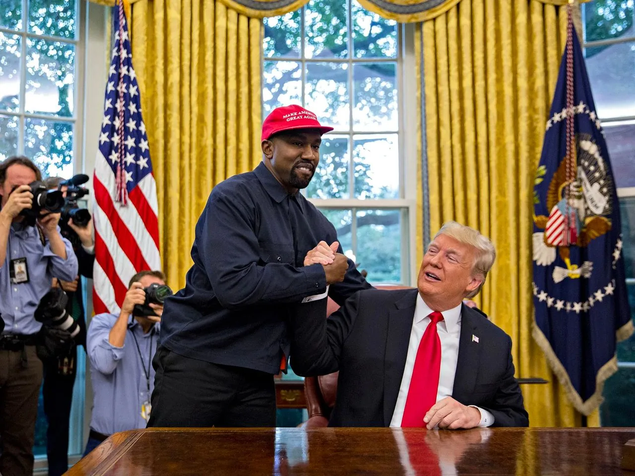 President Trump is seated in the Oval Office, which is characterized by its iconic design elements such as the American flag and the presidential seal. The room is lit with natural light filtering through the large windows, highlighting the golden curtains and the wooden desk. Standing beside him is a man wearing a red cap with visible white text on it. The man is dressed in a dark long-sleeved shirt and is engaging in a friendly handshake with President Trump, who is seated. Both individuals appear to be in good spirits, with President Trump looking up at the man, smiling. In the background, several photographers are capturing the moment, their cameras aimed towards the handshake. The atmosphere is lively, with a mix of formal and casual elements due to the setting and the interaction between the two individuals.