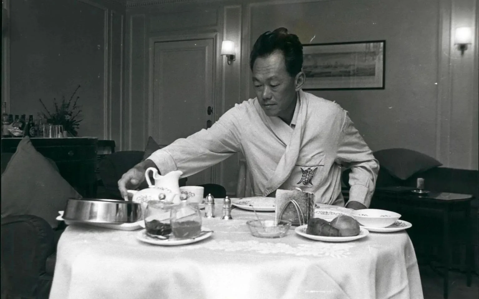 Lee Kuan Yew is depicted in this vintage black-and-white photograph, seated at a table in a hotel room. The setting suggests a moment of quiet leisure, as he is dressed in a robe with a distinctive emblem, indicating possibly a sense of comfort and relaxation. The dining table is elegantly set, showcasing a teapot, cups, and various dishes including bread rolls and what appears to be jam, suggesting a breakfast scene.
Lee Kuan Yew is reaching for a cup or pot, his expression focused and calm. The room's decor is classic with a framed picture on the wall and softly lit lamps, enhancing a serene atmosphere. The background features a sideboard with assorted bottles and glasses, hinting at the amenities of a hotel suite. This image captures a moment of personal routine amidst what could have been a busy schedule, offering a glimpse into the lifestyle and era of Lee Kuan Yew.