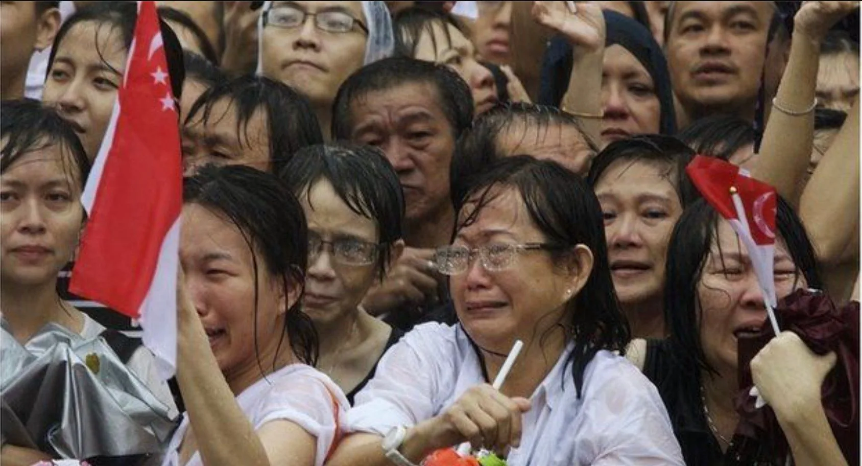 The image captures a poignant scene of emotional intensity, depicting a group of people gathered closely together in the rain. The mood is somber and reflective, with many individuals visibly crying and appearing deeply moved. The people are holding small flags of Singapore, indicating a national event or moment of mourning. The scene is set outdoors, and rainwater glistens on the faces and glasses of those present, adding to the atmosphere of solemnity. The tightly packed crowd suggests a significant turnout, with individuals dressed mostly in casual attire that has become soaked from the rain. The expressions on the faces range from grief-stricken to contemplative, highlighting the collective emotion shared among the attendees. The overall composition suggests a unified display of national sentiment, possibly during a memorial or tribute event. The lighting is natural, softened by the overcast sky, and the colors are muted, with the bright red of the Singapore flag standing out against the otherwise subdued palette.