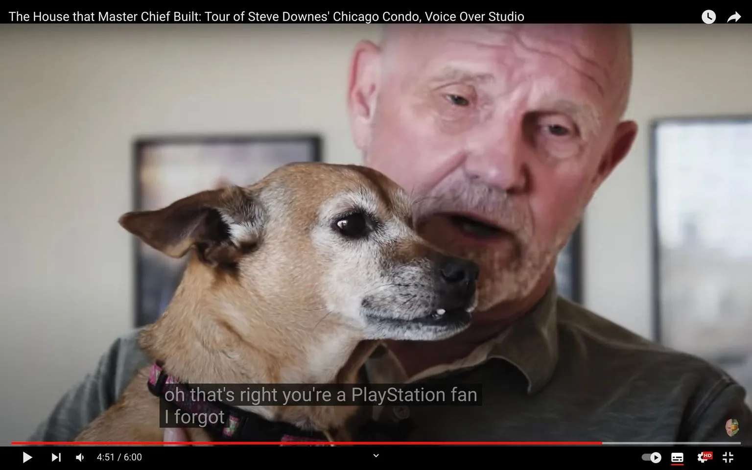 The image is a frame from a YouTube video titled "The House that Master Chief Built: Tour of Steve Downes' Chicago Condo, Voice Over Studio." It features a close-up of a man, identified by the hint as Steve Downes, holding a small dog. The man has a bald head, facial hair, and is wearing a collared shirt, suggesting a casual and relaxed setting. He appears to be speaking, as indicated by the subtitles that read, "oh that's right you're a PlayStation fan I forgot," adding a humorous touch to the scene. The dog, possibly a small mixed breed with light brown fur, is looking attentively ahead. The background is slightly blurred, indicating a focus on the man and the dog. The lighting is soft and evenly distributed, typical of an indoor interview or personal tour video. The video likely explores aspects of Steve Downes' home and professional life, particularly his voice acting career related to the character of Master Chief.
- Title: "The House that Master Chief Built: Tour of Steve Downes' Chicago Condo, Voice Over Studio"
- Site / app: "YouTube"
- Captions / subtitles: "oh that's right you're a PlayStation fan I forgot"