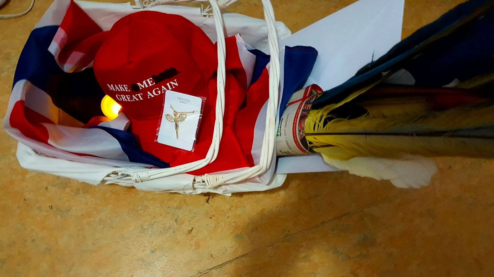 The image depicts a basket containing a variety of items placed on a floor. Central to the composition is a red hat with the text "MAKE ME GREAT AGAIN" embroidered in white. The hat is partially covered by a red, white, and blue fabric, possibly a flag. Resting on the hat is a card displaying a gold-colored necklace labeled "CRYSTAL" in capital letters. Next to these items, there is a set of colorful feathers wrapped in paper. The lighting suggests an indoor setting, with a warm, ambient glow casting soft shadows. The basket is white and appears to be woven, adding a rustic touch to the scene. The mix of items suggests a themed collection or a gift arrangement, with political and decorative elements combined.