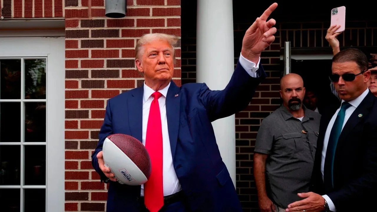 President Trump is depicted in this image standing outdoors in front of a brick building. He is holding a football in his left hand and gesturing with his right hand, pointing upwards. The football has the printed text "TRUMP 2024" on it. President Trump is wearing a dark blue suit with a white shirt and a bright red tie, which stands out against the background. His expression appears confident and focused. To the right of President Trump, a man in sunglasses and a suit, possibly a security personnel, is present, reinforcing the sense of a public event. Behind them, a man in a gray shirt stands near the doorway, adding to the atmosphere of security and attention. The lighting suggests it is daytime, and the presence of a smartphone being held up in the background indicates that people are capturing the moment. The scene conveys a political ambiance, with the football symbolizing a campaign or event-related theme.