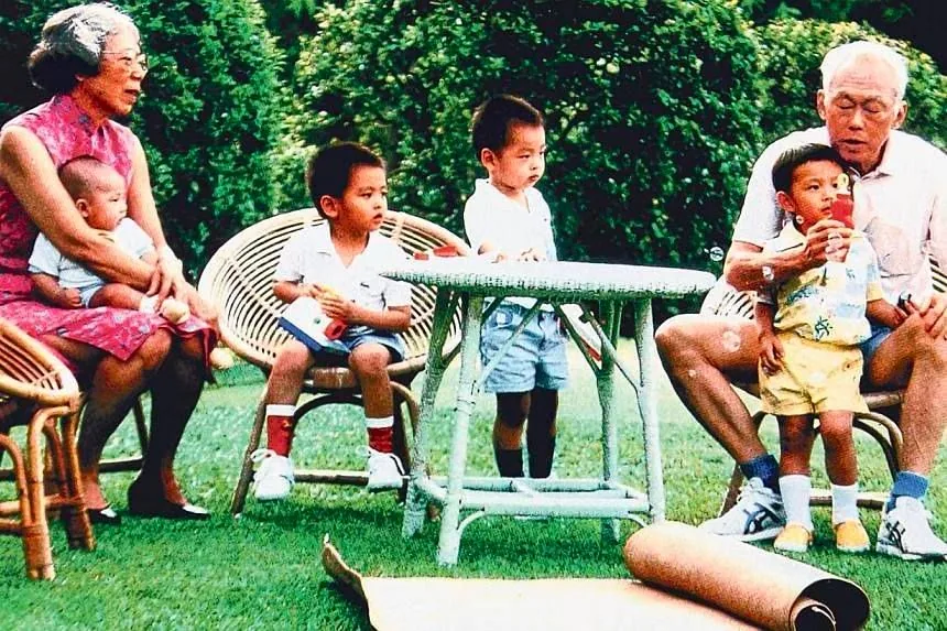 Lee Kuan Yew is depicted in this outdoor family scene, sitting on a wicker chair with a small child on his lap. The setting is a lush garden, with green grass and bushes in the background, creating a peaceful and natural atmosphere. He is wearing a light-colored polo shirt and shorts, exuding a relaxed demeanor as he interacts with the child, possibly his grandchild, who is holding a red object, perhaps a toy or snack.
Beside them, a woman is seated with another child on her lap. She is wearing a traditional sleeveless dress with a floral pattern. The children around the table are dressed casually, with shorts and shirts, contributing to the informal, leisurely family gathering. The wicker furniture and a small coffee table with a greenish hue add to the cozy and inviting ambiance of the scene. The overall mood is familial and serene, capturing a moment of familial bonding and enjoyment in a garden setting.