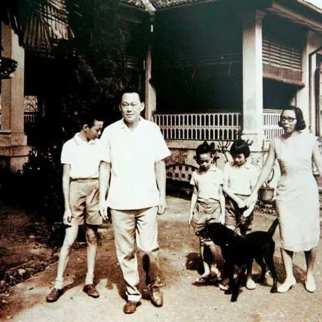 In this vintage photograph, Lee Kuan Yew is seen with his family, walking outside what appears to be a traditional style house. The image captures a moment from the past, featuring Lee Kuan Yew in the center, dressed in a light-colored short-sleeve shirt and trousers, exuding a relaxed but authoritative presence. To his left, a young boy in shorts and a light shirt walks alongside him, while on his right, a woman in a simple, elegant dress holds the leash of a small black dog.
The background shows a building with a tiled roof and shaded windows, typical of mid-20th-century architecture. The overall mood of the photograph is serene and candid, reflecting a family outing. The lighting is natural, indicating it was taken during the day. The black-and-white tone of the image adds a timeless quality, emphasizing the historical context of the scene. The family appears engaged in each other’s company, unaware of the camera, capturing an intimate moment of everyday life.