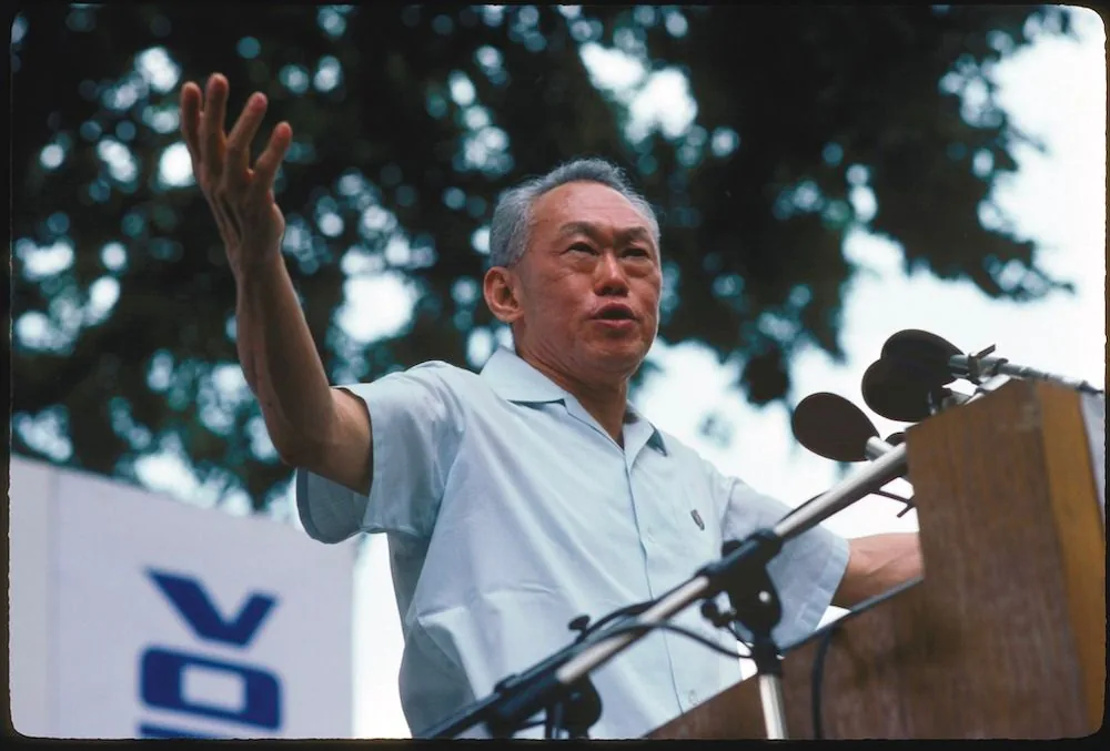 In the image, Lee Kuan Yew is giving an outdoor speech, standing behind a podium equipped with multiple microphones. He is wearing a light-colored short-sleeved shirt, which contrasts with his silver hair, providing a sense of authority and presence. His arms are outstretched in an expressive manner, suggesting he is emphasizing a point during his address. The background is slightly blurred, with the leafy branches of a tree visible, indicating the event is taking place in a natural, open-air setting. The lighting is bright and natural, typical of a daytime event. Lee Kuan Yew's facial expression appears serious and focused, conveying the weight and importance of his message. There is no visible on-screen text or specific branding in the image, allowing the focus to remain on his commanding presence and oratory gesture.