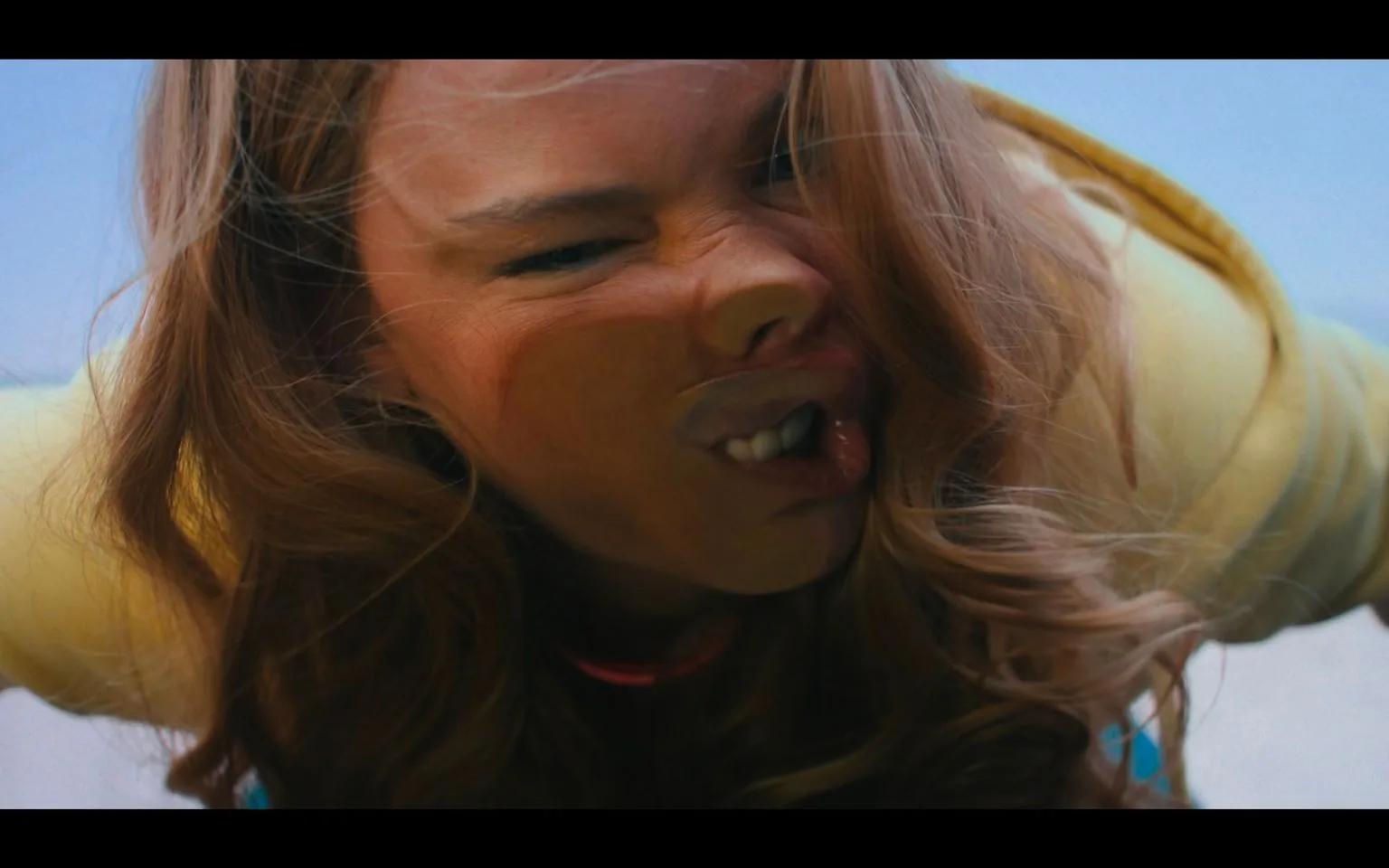 The image is a close-up of a person's face, with their features humorously distorted as if pressed against glass. The person's expression is exaggerated, with a scrunched nose and mouth, as if they are playfully squishing their face. Their eyes are partially closed, adding to the comedic effect. The person has long, wavy hair that frames their face and a yellow garment can be seen, suggesting casual attire, possibly a jacket or sweater. The background is a soft blue, indicating an outdoor setting during the daytime. The overall mood is lighthearted and humorous, capturing a moment of playful expression. The lighting is natural, enhancing the colors in the image.