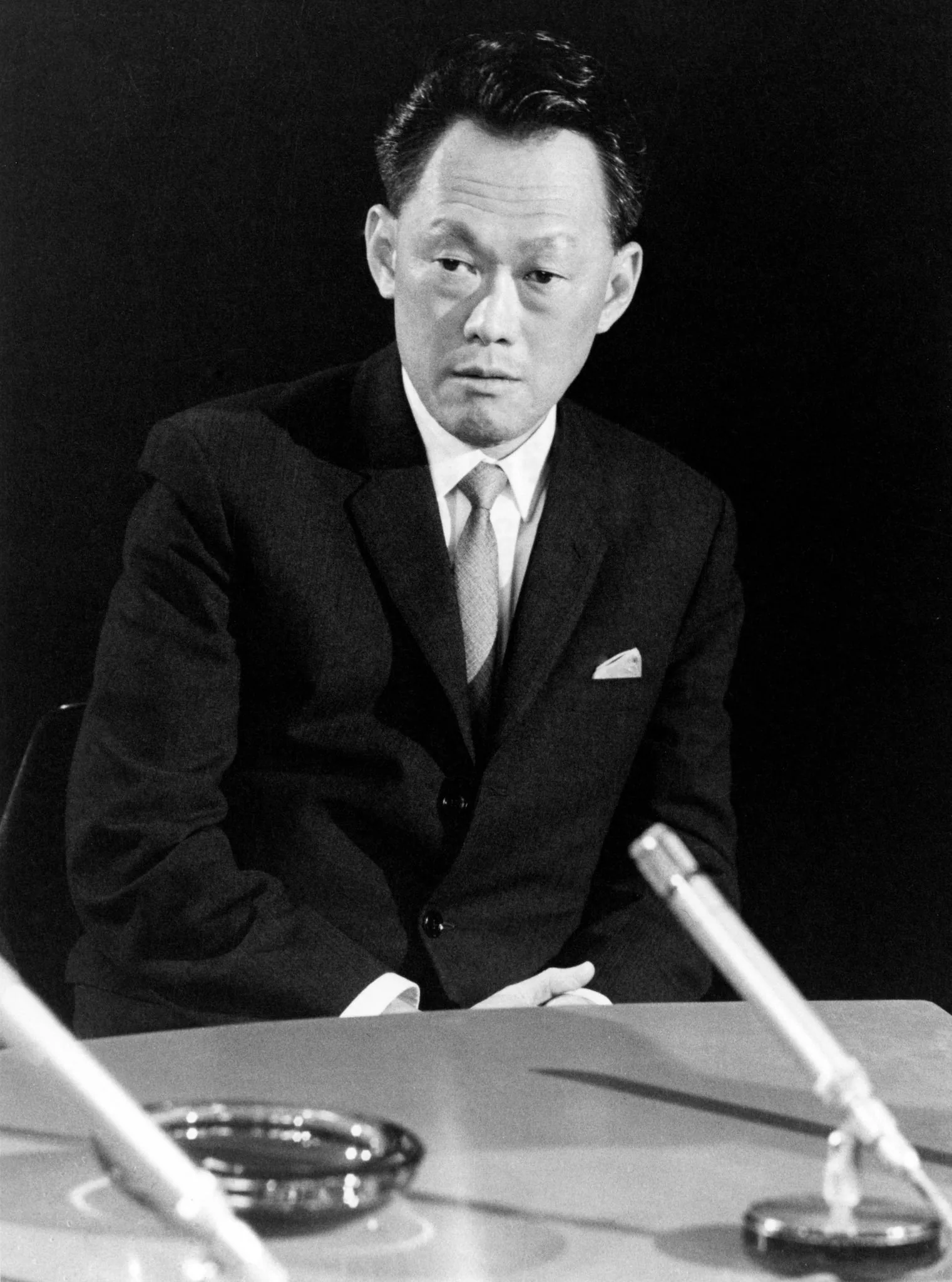 Lee Kuan Yew is captured in a black and white photograph, sitting at a table during what appears to be an interview or a formal discussion. He is dressed in a dark suit with a neatly folded pocket square and a light-colored tie, exuding a sense of professionalism and authority. His hair is neatly combed back, and he wears a serious expression, indicative of deep thought or focus.
The setting is simple and formal, with a dark background that contrasts with his lighter attire, highlighting his presence. In the foreground, there is a microphone visible, suggesting a broadcast or recorded interview. The tabletop is sparsely occupied, with what seems to be an ashtray near the microphone, indicative of the era when the photograph was likely taken. The composition centers on Lee Kuan Yew, emphasizing his significance in the scene. The overall mood conveys an atmosphere of importance and gravity, fitting for a political figure engaged in a public or significant conversation.