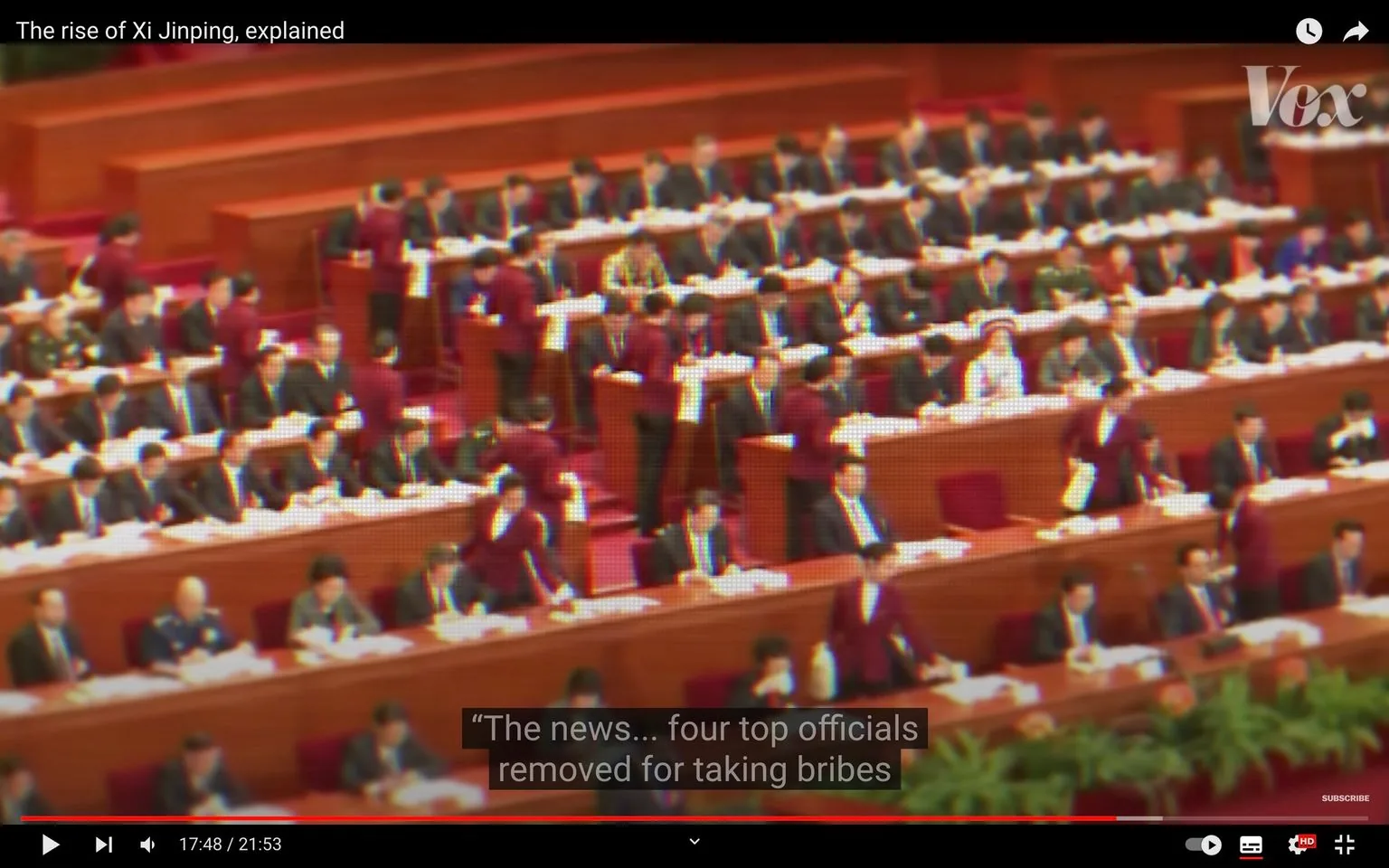 ```
This video frame from a Vox YouTube video titled "The rise of Xi Jinping, explained" depicts a formal governmental assembly, likely within a Chinese political context. The setting is a large, well-lit auditorium with rows of long wooden desks occupied by numerous attendees in suits, indicative of a serious political meeting or session. The scene captures a moment of significant activity with several attendants wearing maroon uniforms moving between the rows, distributing or collecting documents. The mood is formal and structured, reflecting the procedural nature of such gatherings.
In the foreground and background, individuals are seated in orderly rows, focused on paperwork or speaking with colleagues, suggesting engagement with official proceedings. The color palette is dominated by the rich reds and browns of the seating and desks, complemented by the dark suits of the participants, creating a cohesive and serious visual tone. The subtitles highlight a moment of political significance, referencing the removal of top officials for bribery, adding a layer of narrative tension to the scene. The presence of plants in the front suggests an attempt to soften the otherwise stern environment, typical of formal governmental settings.