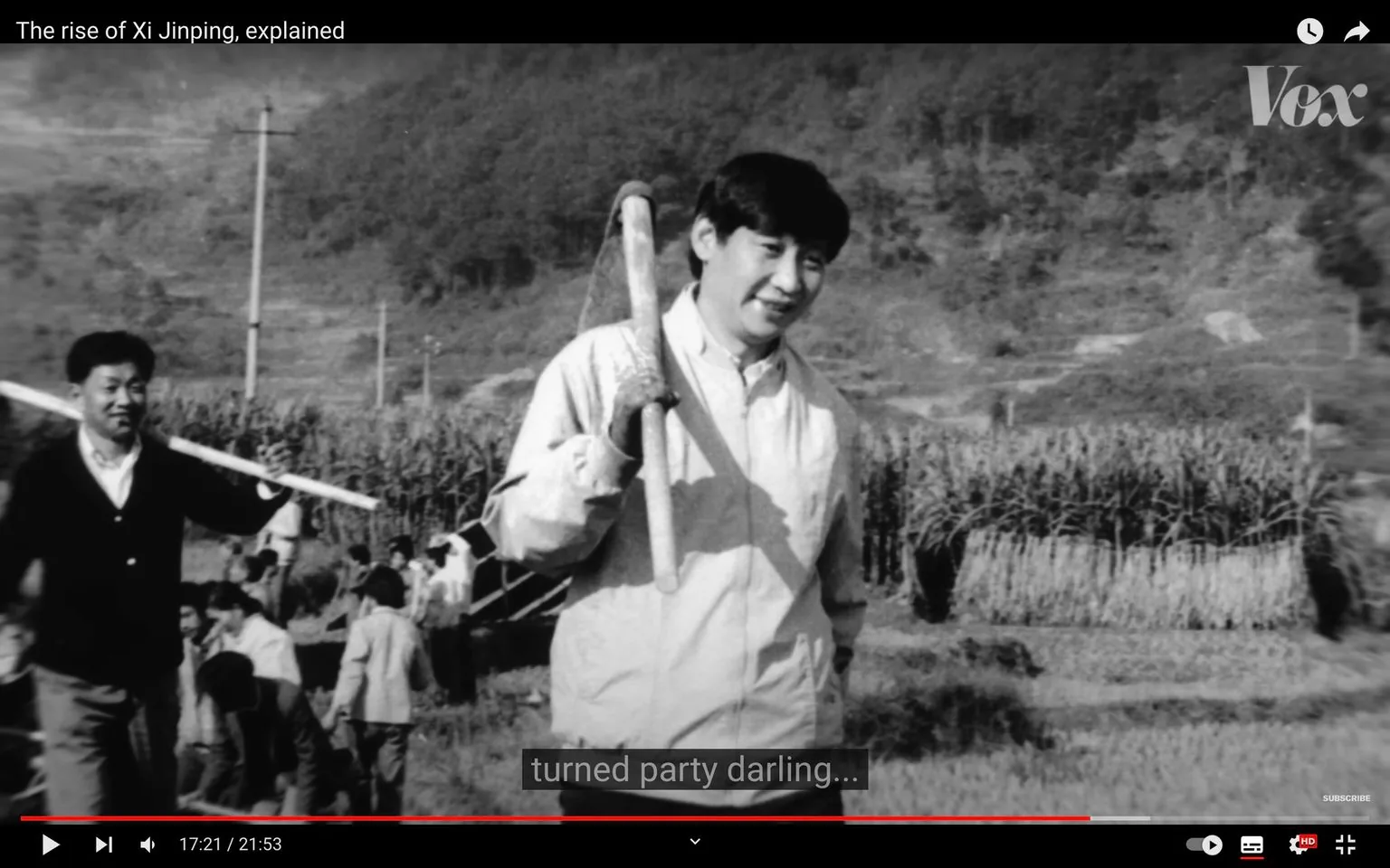 The black-and-white image depicts a young Xi Jinping, who appears to be standing outdoors in a rural setting. He is holding a long-handled tool over his shoulder, suggesting a farming or labor activity. His attire is casual, consisting of a light-colored jacket and trousers, appropriate for outdoor work. The background features a field with tall grasses or crops and a few other people engaged in similar activities, creating a sense of community or group effort.
The overall mood is one of diligence and simplicity, with Young Xi Jinping smiling slightly, exuding a sense of determination and approachability. The image is framed with a focus on him in the foreground, making him the central figure of interest. The natural landscape, with hills and vegetation, provides a serene backdrop that emphasizes his connection to the land and work.
The video frame is from a Vox video titled "The rise of Xi Jinping, explained," and the visible subtitle reads, "turned party darling..." This suggests a narrative of transformation and political ascent. The composition captures a significant moment in his early life, reflecting his humble beginnings and subsequent rise in Chinese politics.