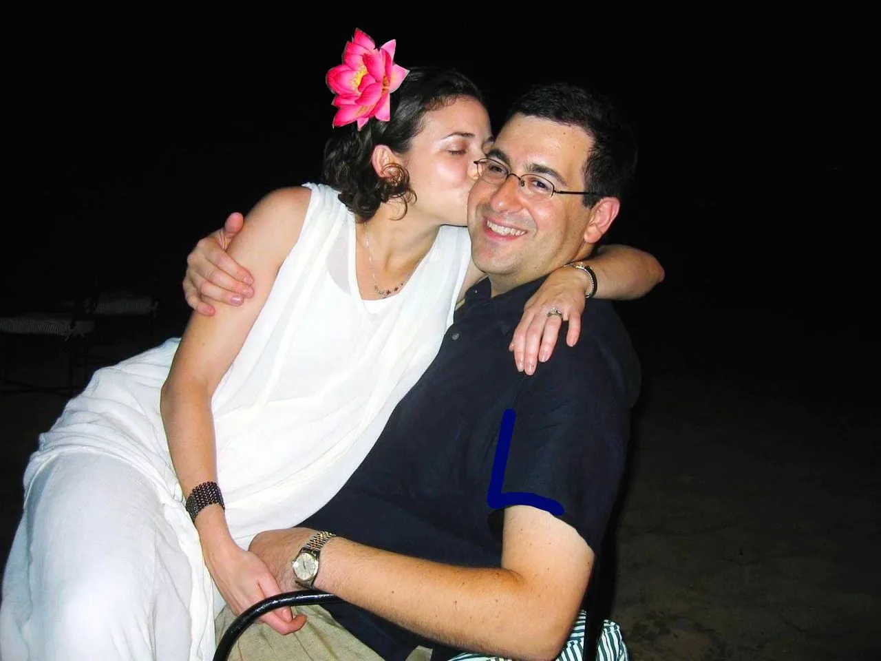 Sheryl Sandberg and Dave Goldberg are depicted in this image. It's a nighttime scene with a dark background, suggesting an outdoor setting. Sheryl Sandberg is wearing a white outfit and has a large pink flower in her hair, adding a vibrant touch. She is affectionately kissing Dave Goldberg on the cheek, who is seated and smiling warmly. Dave is wearing glasses and a dark shirt. The two are sharing a tender moment, and their expressions convey happiness and contentment. The lighting is soft, focusing primarily on them, creating an intimate and relaxed atmosphere. Both display watches on their wrists, hinting at their style. The overall composition highlights a personal and joyful interaction between the two individuals.