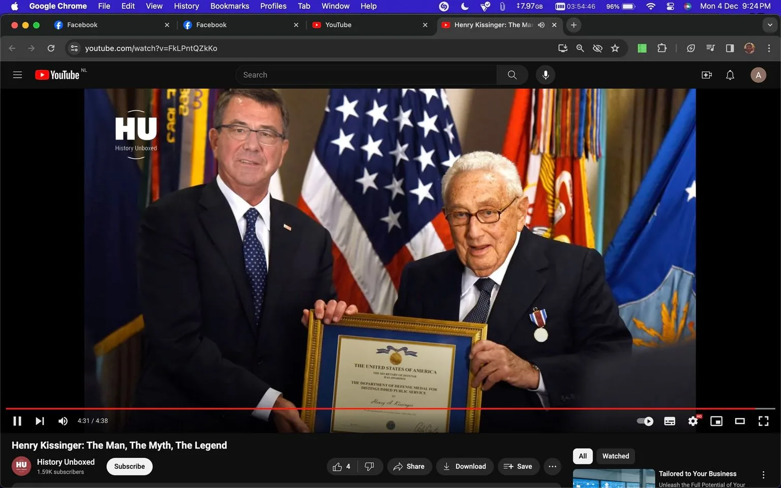 Henry Kissinger is depicted in a formal award ceremony setting, where he is receiving a framed certificate. The certificate prominently displays text related to the United States of America and the Department of Defense Medal for Distinguished Public Service, with Henry A. Kissinger's name on it. He is wearing a dark suit with a tie, and a medal is pinned to his lapel, indicating an honorary event. Beside him stands a man in a similar formal suit, likely a presenter or official, holding the frame with Henry Kissinger. The background features American flags and other military banners, reinforcing the official and ceremonial nature of the event. The lighting is bright and clear, suitable for a formal indoor occasion. The video is hosted on YouTube, titled "Henry Kissinger: The Man, The Myth, The Legend," and is presented by the channel History Unboxed. The screenshot captures a moment towards the end of the video at the 4:31 mark.